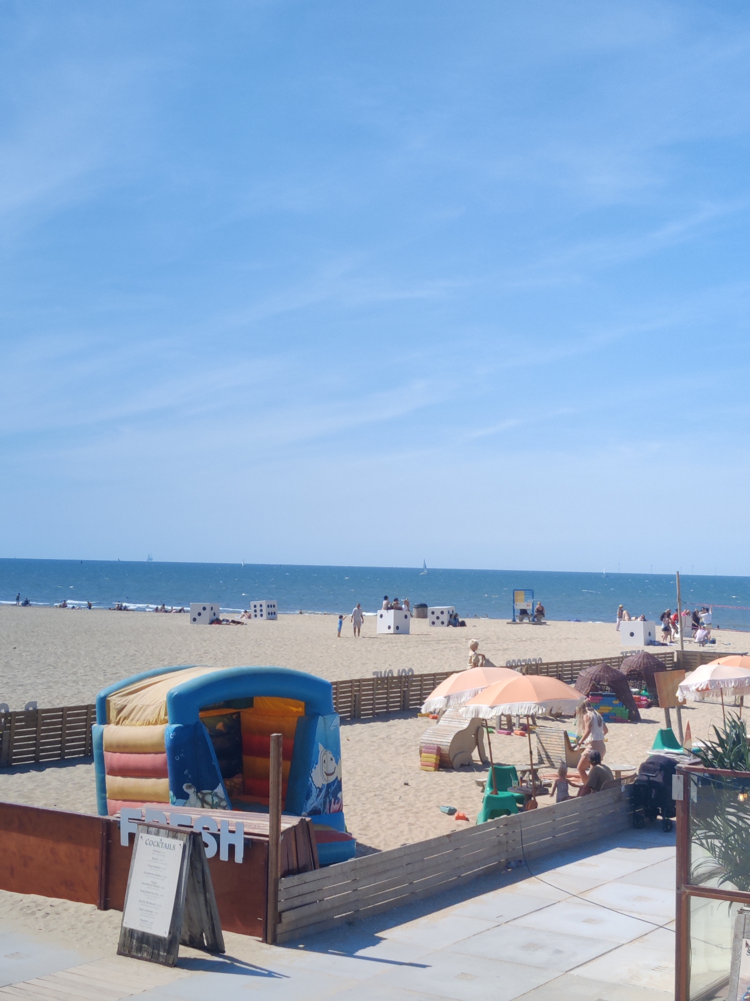 This image depicts a sunny day at a beach in The Hague, Netherlands. The scene is crowded with people enjoying the sand and sea.
In the foreground, a wooden barrier encloses an area on the sand with a blue and yellow inflatable play structure featuring cartoon characters. Next to it, a wooden stand displays a menu titled "COCKTAILS". The beach is scattered with large white cubes, some with black dots, resembling dice. Beach umbrellas and small tents are set up, providing shade for beachgoers.
Further in the background, people are seen sunbathing on the sand, playing near the water's edge, and walking along the shore. Sailboats are visible on the horizon. The sky is clear and blue, indicating pleasant weather. The overall impression is of a lively and family-friendly beach environment.