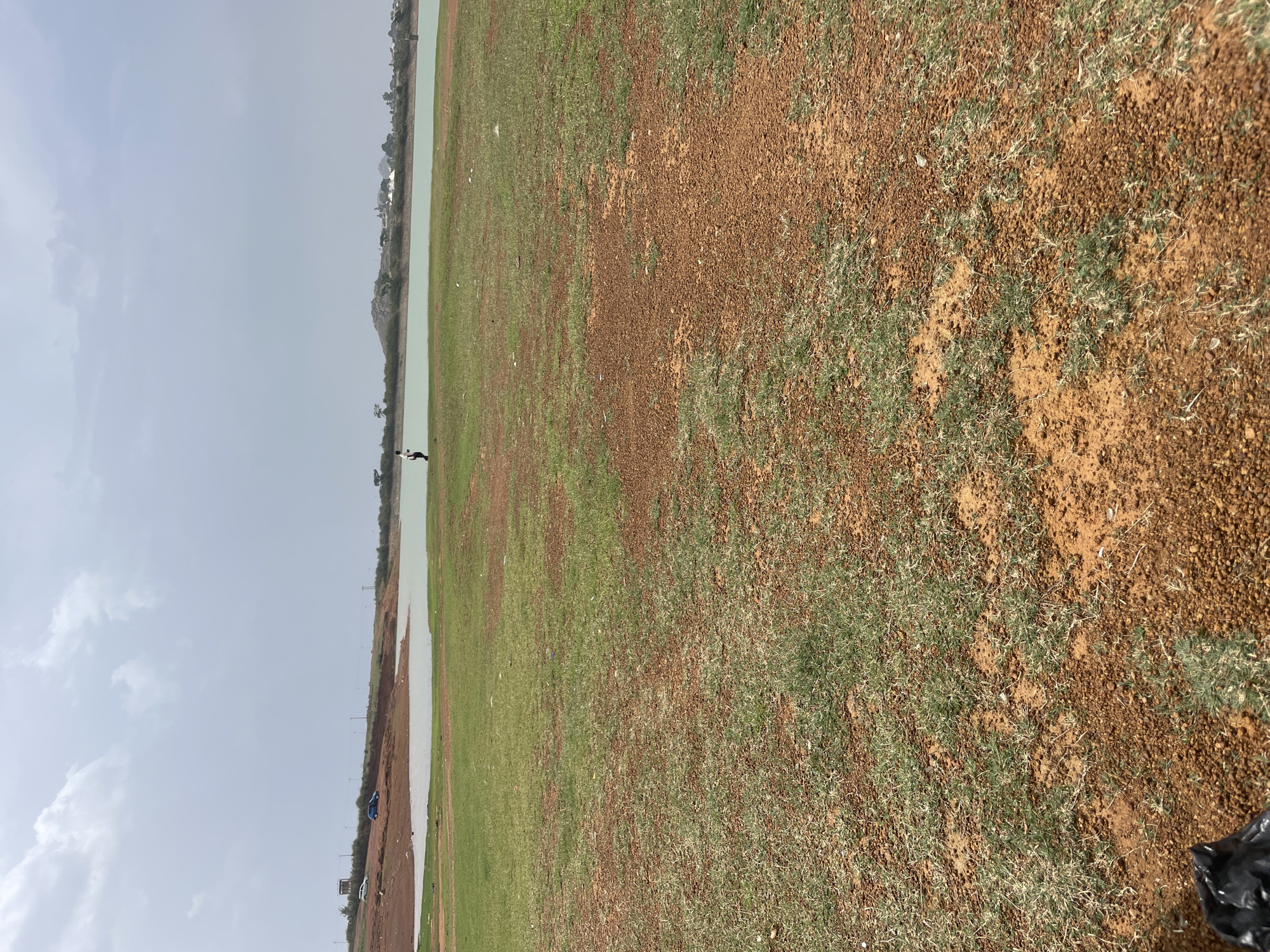 The image depicts a landscape near Nabatum, Nigeria. A body of water runs horizontally across the left side of the frame, with buildings and trees visible on the far bank. A single, unidentifiable person stands near the water's edge. The foreground is characterized by red-brown soil interspersed with patches of green grass, extending across the majority of the scene. The sky is overcast. A black plastic bag is partially visible in the lower right corner of the frame.