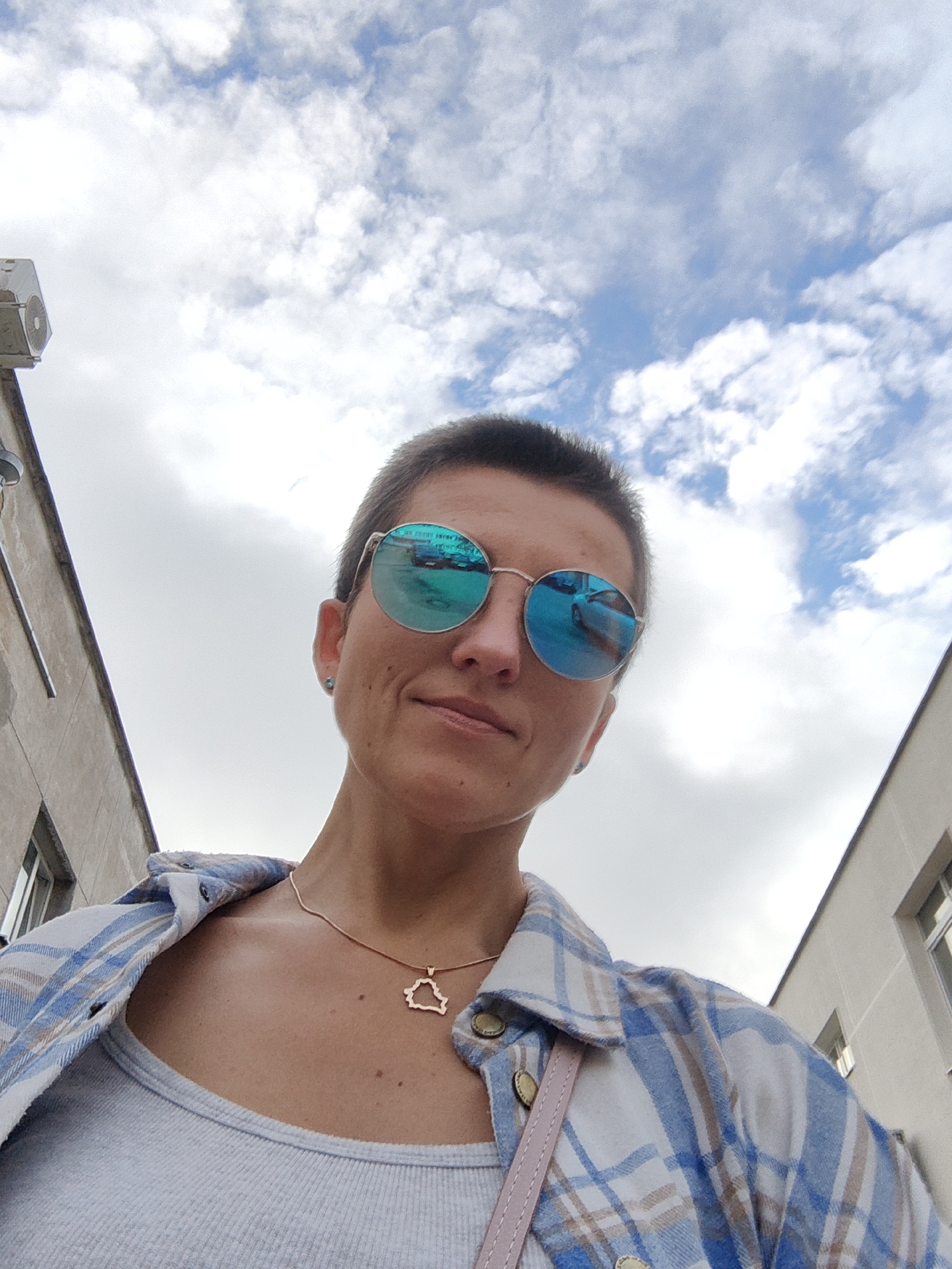 A low-angle, close-up, outdoor selfie shows a person with very short hair and round, blue-tinted sunglasses. The person is wearing a light-colored tank top under an open, blue and white plaid shirt. A thin gold chain with a pendant shaped like the outline of Belarus is visible around the person's neck. The background consists of a cloudy sky and the sides of two buildings, suggesting an urban environment. The reflection in the sunglasses shows what appear to be cars and possibly buildings. The image is likely taken in Minsk, Belarus, given the pendant.