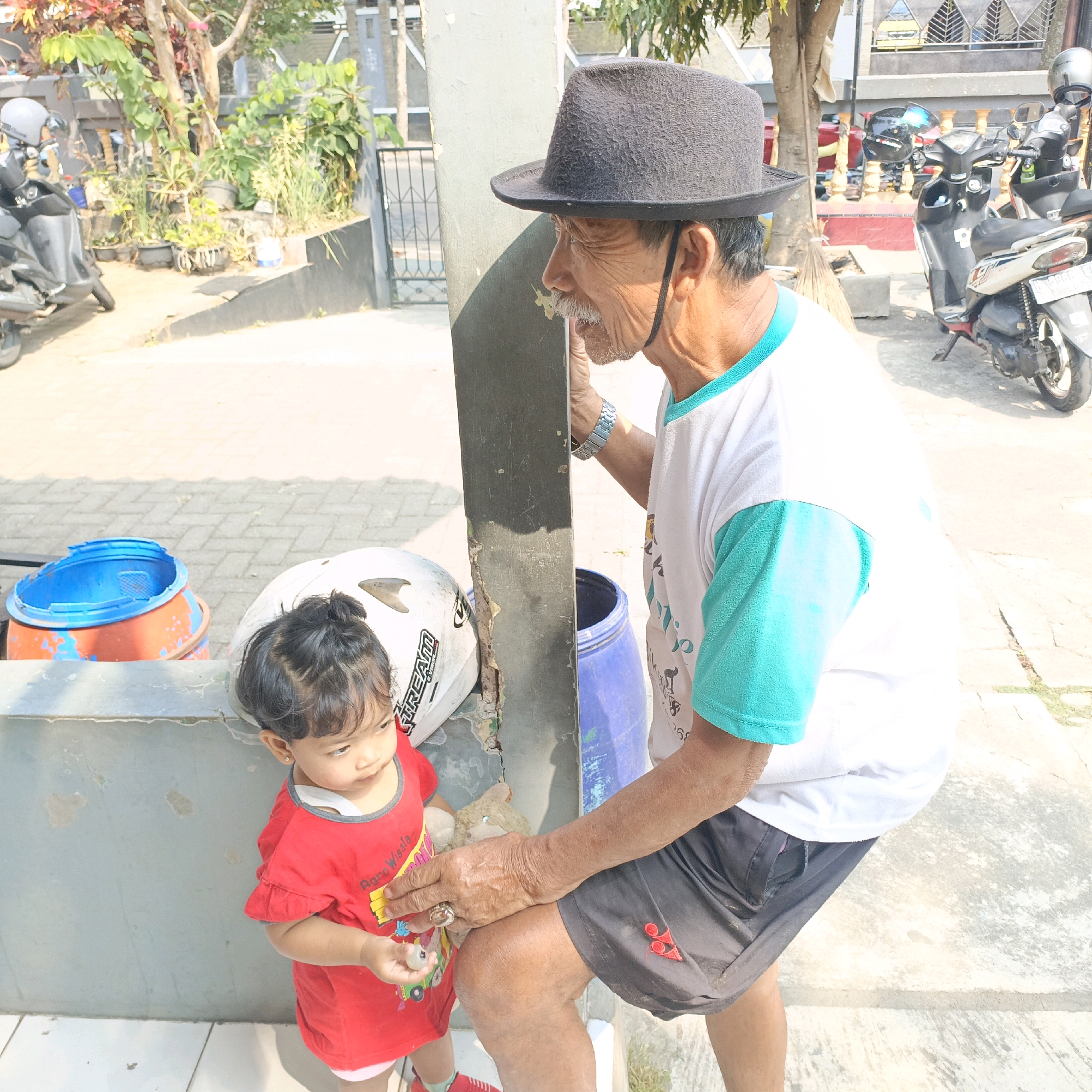 An elderly Indonesian man wearing a dark grey fedora and a white t-shirt with teal sleeves stands to the right of a dark grey pillar, looking down and to his left at a young Indonesian girl. The man has weathered skin, a mustache, and is wearing a watch on his left wrist and a ring on his right index finger. The girl, to the left of the pillar, is wearing a red t-shirt and is looking down and to her right. She is holding a small, light-colored object in her right hand. To the left of the girl is a white helmet. Behind the man and to the right is a blue plastic barrel. In the background, there are parked motorcycles and vegetation. The scene appears to be outdoors, possibly in front of a building or house, on a sunny day. The location is identified as Salatiga, Indonesia.