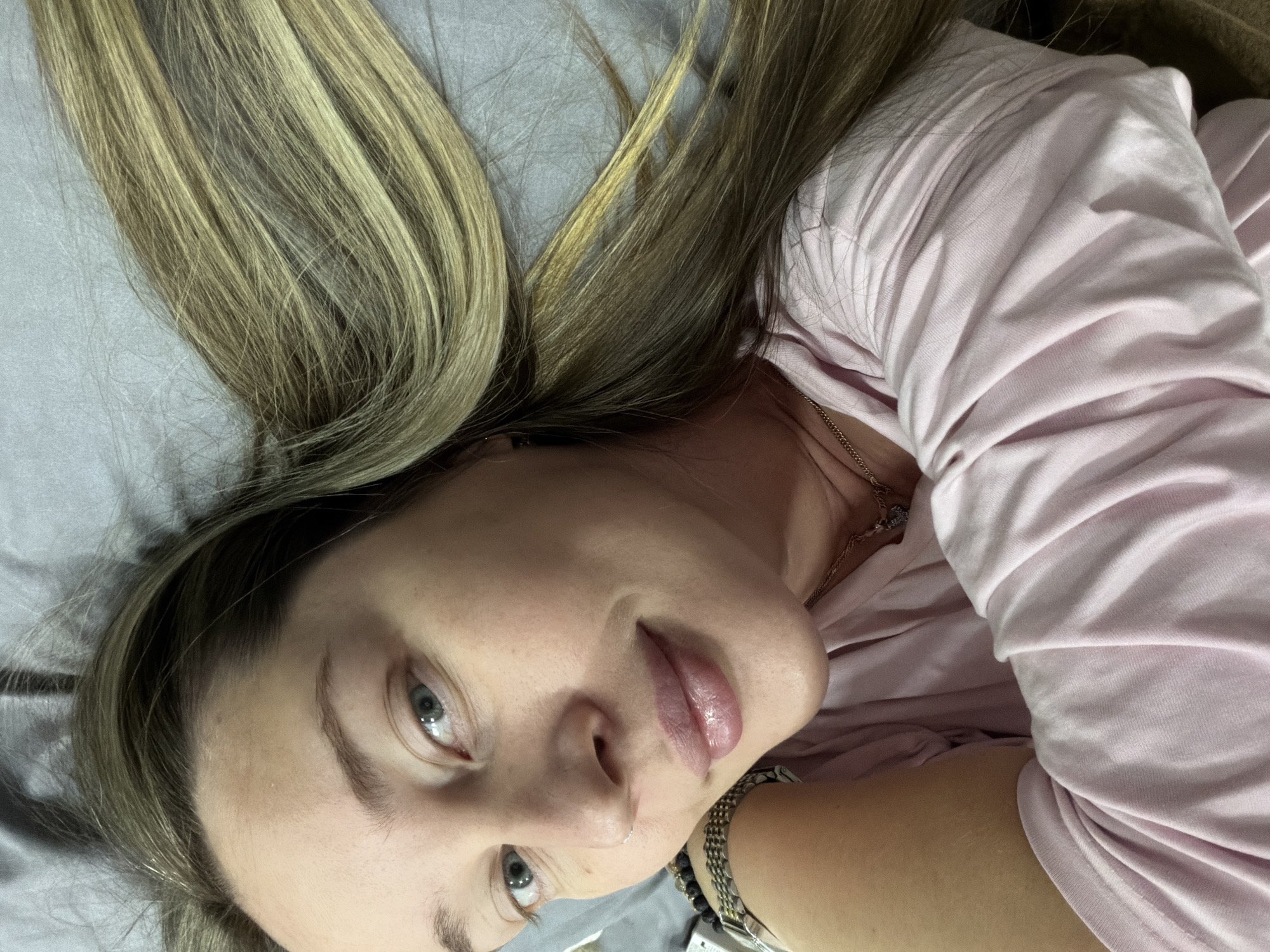 A woman is lying on a gray surface, presumably a bed, with her blonde hair spread out around her head. She is wearing a pink top. She has light skin, blue eyes, and is looking up toward the camera. A chain necklace is visible on her neck. The location in Chaikovsky, Russia, cannot be determined from this image.