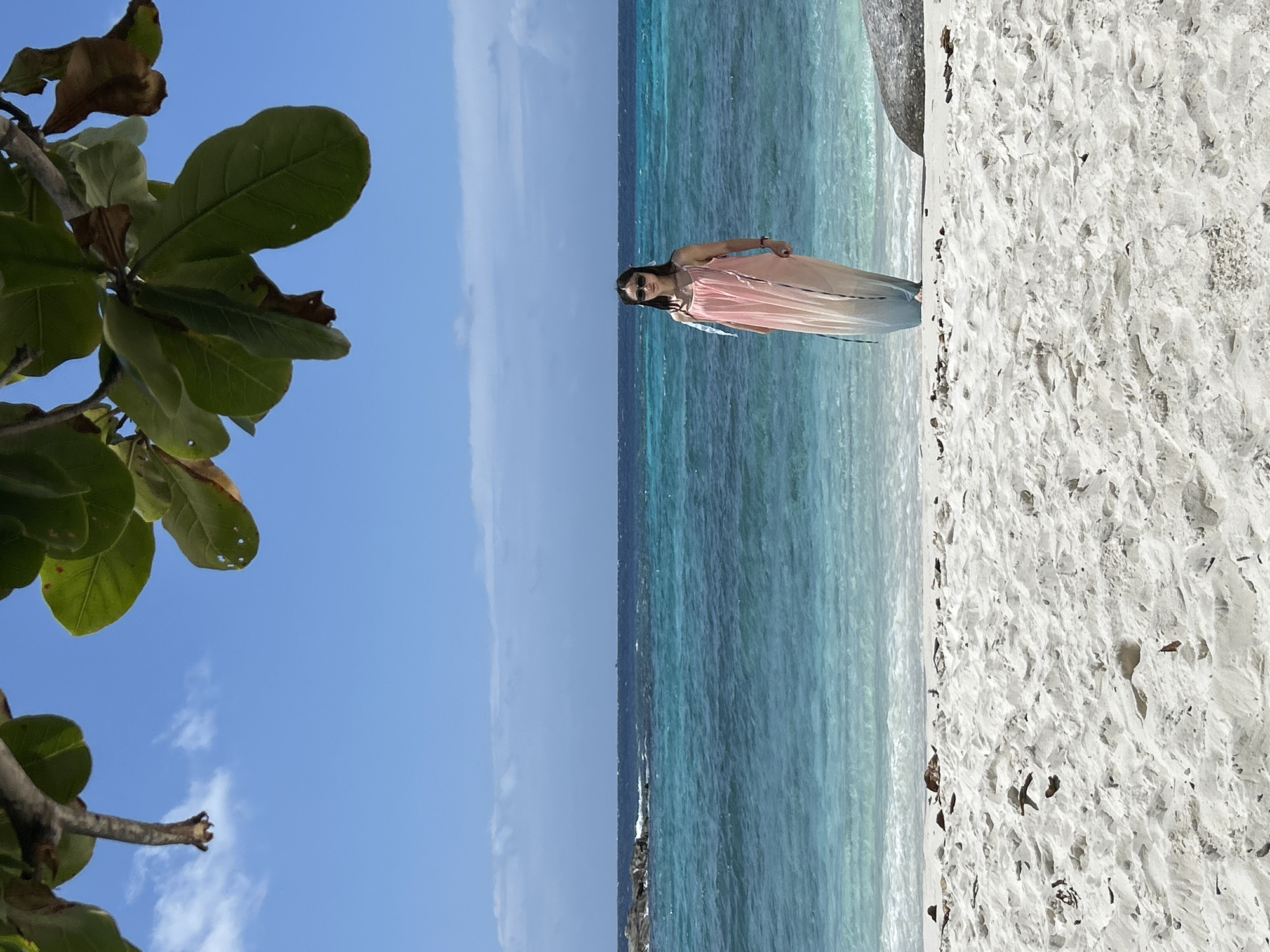 A woman stands on a sandy beach in Rawai, Thailand, facing the camera. She is wearing a long, light pink and white ombre dress, sunglasses, and her hair is pulled back. Behind her, the turquoise ocean stretches to the horizon, meeting a deep blue sky with scattered clouds. The foreground is dominated by white sand with scattered debris. Lush green leaves hang from a branch in the upper left corner of the frame, casting a partial shadow over the scene. The water appears calm with gentle waves lapping the shore.