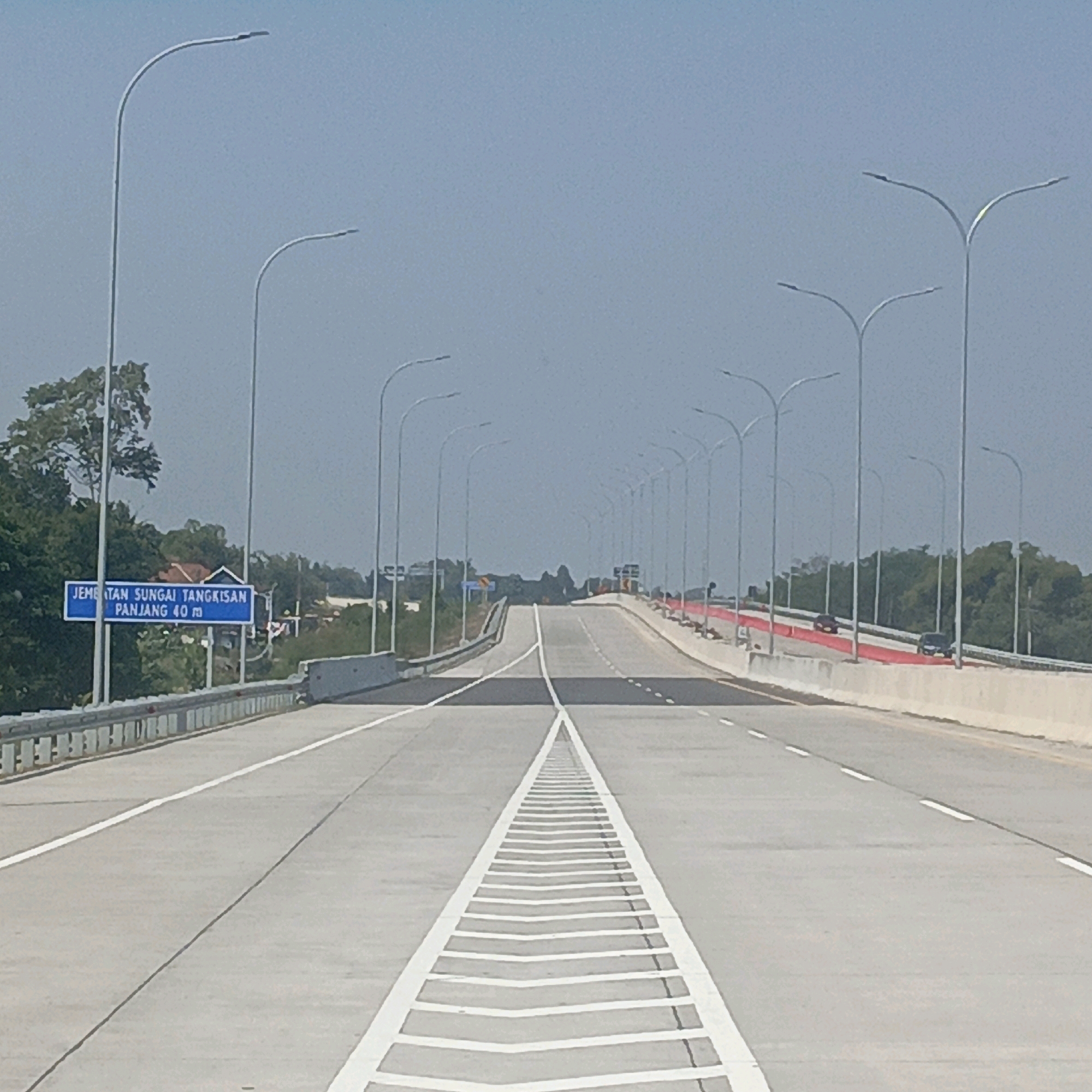 The image depicts a highway with a clear sky and lush green trees on either side. A blue sign on the left side of the highway reads "JEMBATAN SUNGAI TANGKISAN PANJANG 40 m", which translates to "Tangkisan River Bridge Length 40 m". This indicates the location is the Tangkisan River Bridge, measuring 40 meters in length, in the city of Joton, Indonesia. The highway is lined with numerous lampposts and has a well-maintained asphalt surface with clear white lane markings. Two vehicles are visible on the highway, one black and one dark red, traveling in the same direction. The environment suggests a sunny day, with the highway appearing to be newly constructed or recently repaved.