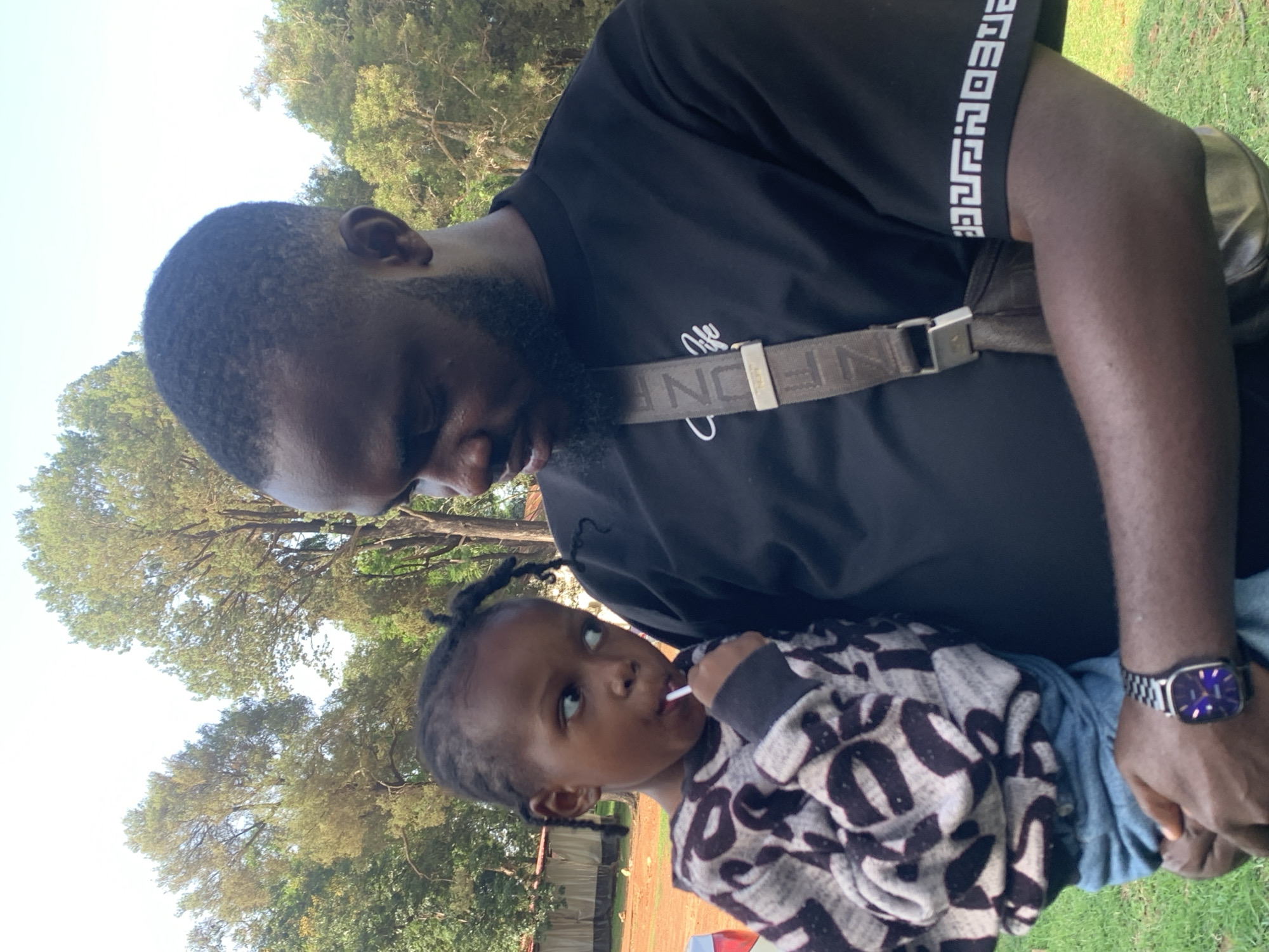 The image shows a dark-skinned man holding a young child outdoors. The man is wearing a black t-shirt with white graphic designs on the sleeves and a crossbody bag. The child is wearing a patterned sweater and jeans, and is holding a white lollipop. The background features green grass, trees, and some buildings, suggesting an outdoor setting in a city or suburban area. The lighting indicates it is daytime, likely with bright sunlight filtering through the trees. The image captures a candid moment between the man and child. The location is identified as the city of Jos, Nigeria, based on the context provided.