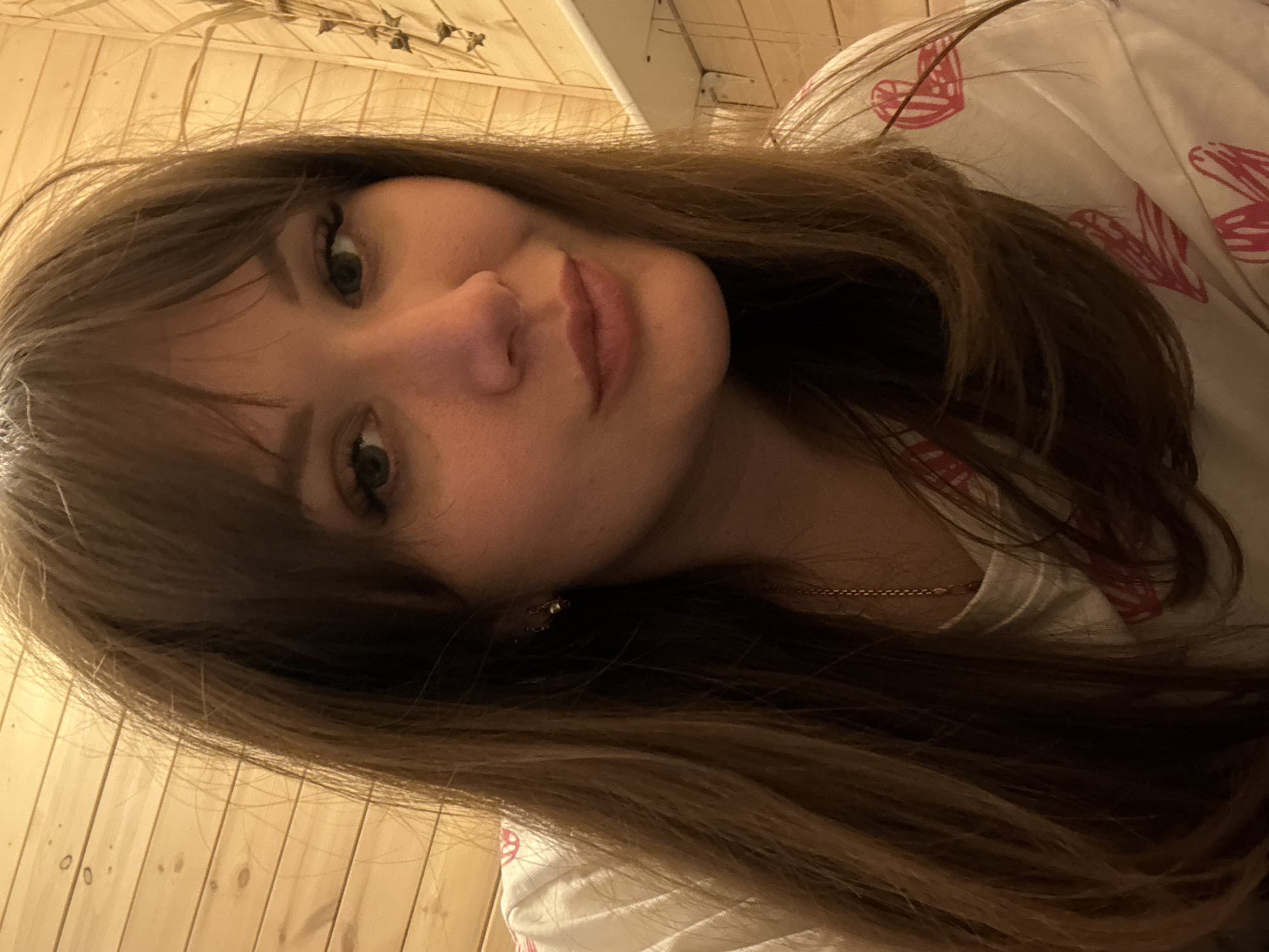 A close-up selfie depicts a woman with light skin, brown hair, and green eyes, wearing a white shirt patterned with pink hearts. She has full lips, a slightly upturned nose, and a fringe of bangs. A gold chain necklace is visible around her neck, and she wears a small earring. The background consists of wooden paneling. The location of the photograph is not evident within the frame, and therefore, the specific location in the city of Taman', Russia cannot be determined.