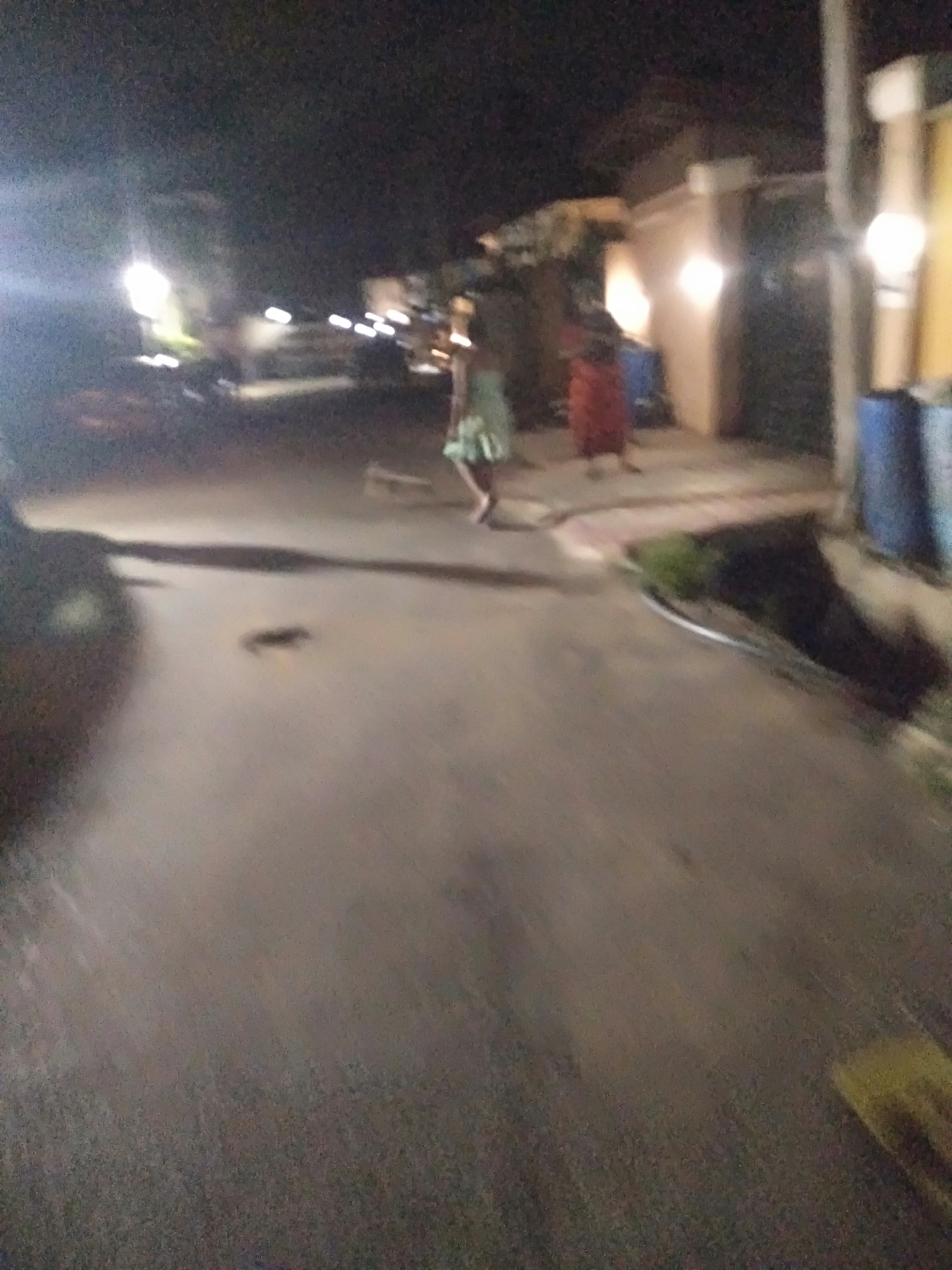 The image shows a dimly lit street at night. Two individuals are visible, one in a green dress walking, and another in a red dress standing near the sidewalk. Several buildings line the street with lights illuminating their fronts. There is a blue trash container to the right. A vehicle is visible in the distance. The location appears to be a residential street.