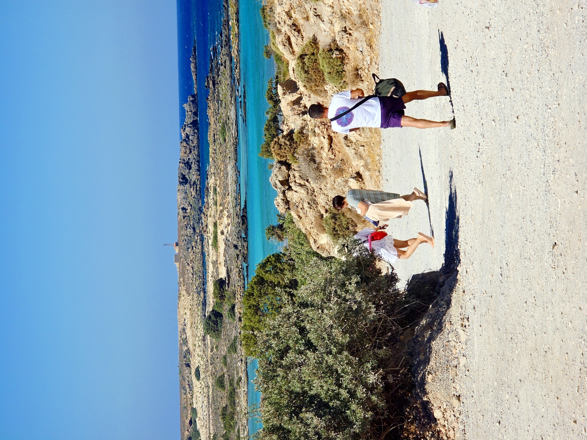 An eye-level, medium shot captures three individuals walking on a dirt path in a coastal area. The foreground is dominated by the light-colored, gravelly path with shadows cast by unseen objects. To the left, a large, dense green bush partially obscures the path.
In the mid-ground, three people are walking away from the camera. The person on the far left is a child wearing a white dress and a red bag. Next to the child is an adult in a light-colored dress, carrying a beige towel. On the right, an adult male wears a white t-shirt with a colorful circular design, purple shorts, and flip-flops, carrying a dark green bag over his left shoulder.
The background reveals a rugged, arid landscape with sparse vegetation and rocky outcrops. Beyond this, a body of bright turquoise water stretches to the horizon, meeting a clear blue sky. A rocky coastline with a small, distant structure on a hill is visible across the water. The scene appears to be a sunny day at a coastal location in Greece.