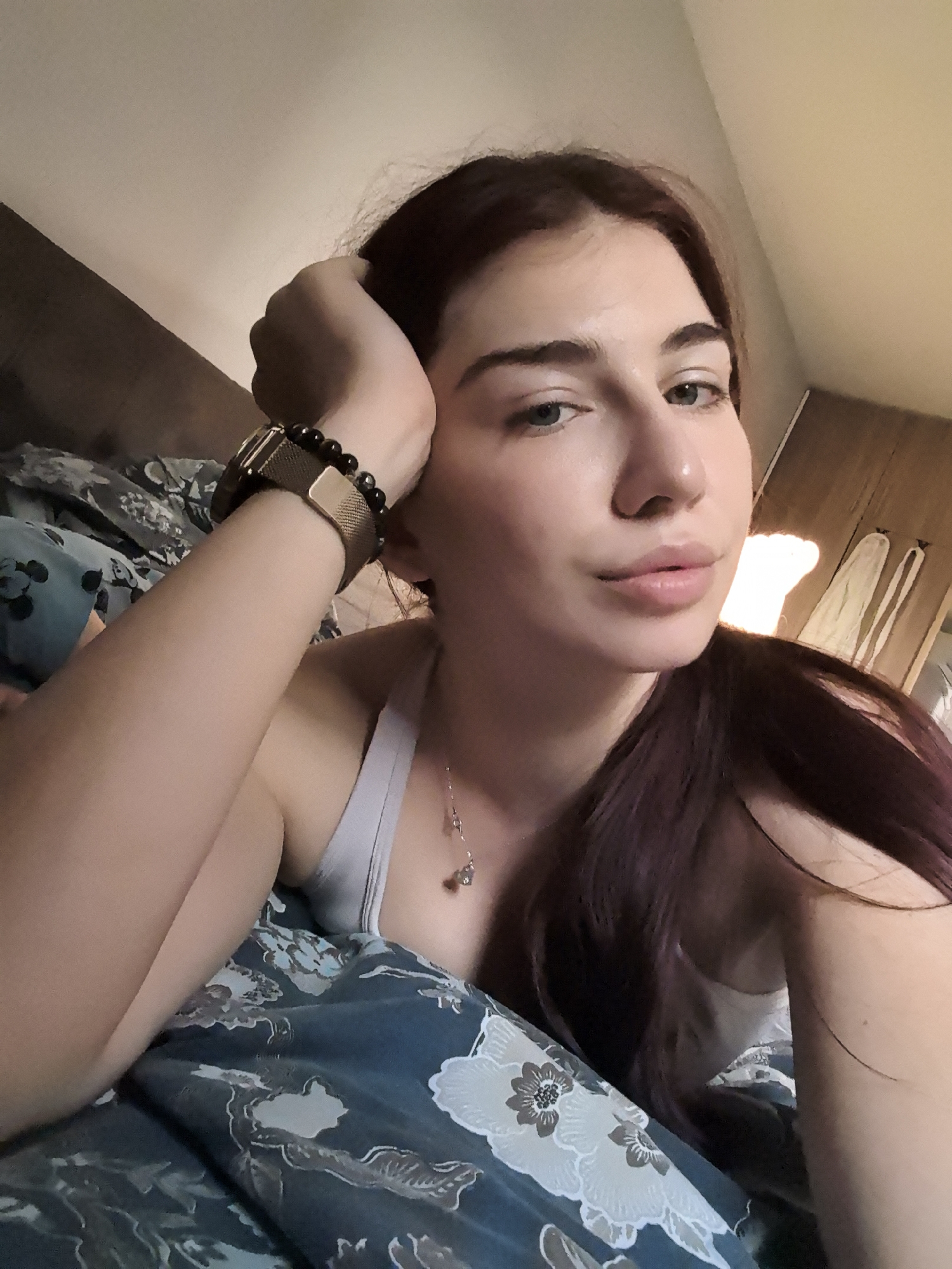 A young woman is captured in a close-up selfie, lying on a bed indoors, likely in Stara Zagora, Bulgaria. She is positioned on her left side, propping her head with her left hand, which gently supports her jawline. Her left arm is visible, wearing a gold-toned watch with a mesh band and a beaded bracelet made of dark, possibly black, stones.
The woman has fair skin, dark reddish-brown hair that falls over her shoulder, and light-colored eyes (appearing greenish-blue). Her eyebrows are natural and dark, complementing her hair color. Her lips are full with a natural, light pink tone. She looks directly at the camera with a calm, neutral, and slightly pensive expression.
She is wearing a light-colored (possibly light grey or white) tank top or camisole, and a delicate silver-colored necklace with a small, light-colored pendant around her neck.
The bed is covered with a dark blue or teal duvet featuring a white and light grey floral pattern. In the background, to the right, a warm, ambient light source casts a glow, illuminating a section of what appears to be a wooden furniture piece or wall panel. Two white or light-colored items, possibly garments or towels, are hanging on a fixture in the background. The lighting suggests an indoor setting with soft, diffused light, possibly during the day or evening. No text is visible in the image.