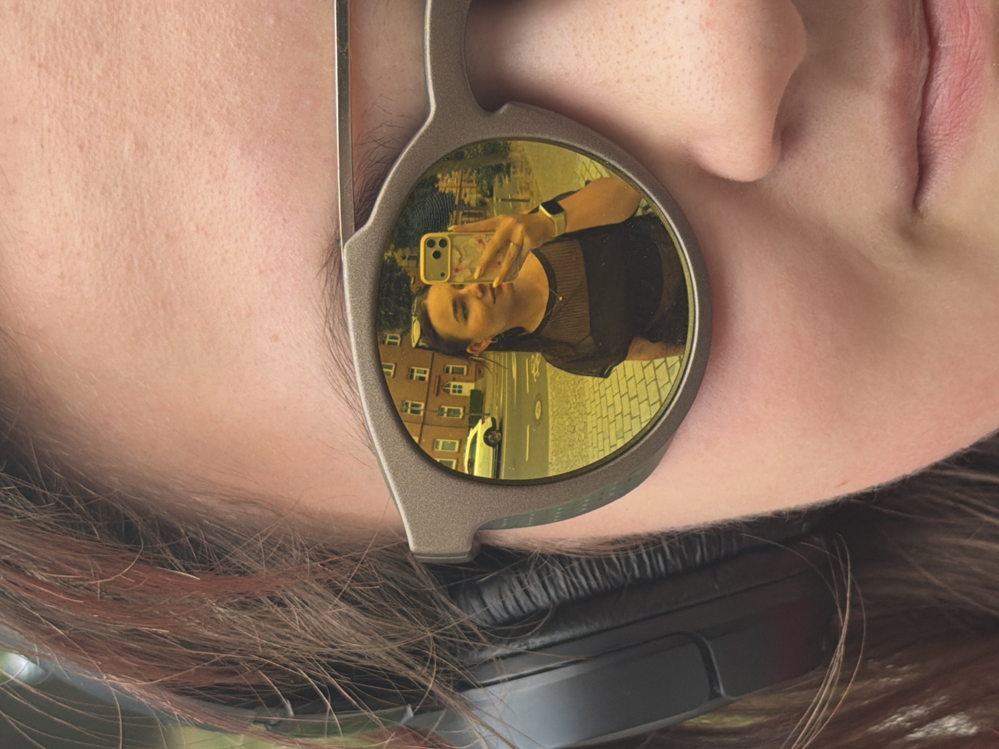 The media file is a close-up, vertically oriented selfie taken by a young person wearing sunglasses with yellow, reflective lenses. The selfie is reflected in the right lens of the sunglasses. The person is holding a smartphone with a floral case to capture the reflection. They are wearing a dark, mesh-like top and a smartwatch on their left wrist. The background visible in the reflection appears to be a street scene with buildings and a parked car. The environment suggests an urban setting during daylight. The overall mood is casual and self-aware, typical of a selfie.
Based on the provided location context of Erfurt, Germany, the buildings in the reflection could potentially be characteristic of architecture found in the city. The paved road and sidewalk also contribute to the urban setting. The lighting suggests it is daytime, and there are no clear indicators of specific weather conditions. The image focuses on the reflection, offering a glimpse of the person's face, shoulders, and the surrounding environment. The details in the reflection, such as the architecture and the street, are not sharp enough to definitively identify a specific landmark in Erfurt.