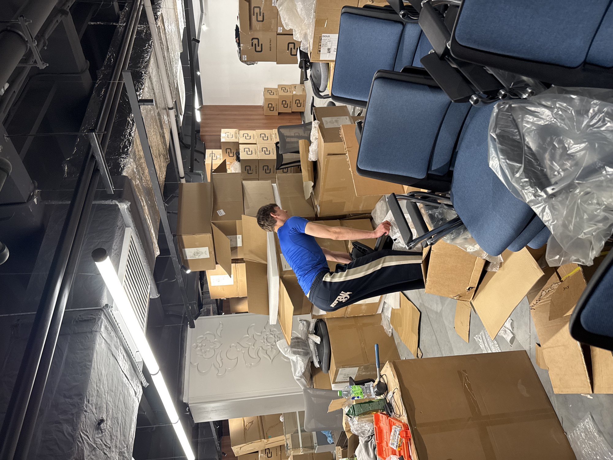 A man in a blue shirt and dark sweatpants sits on a chair amidst numerous cardboard boxes and office chairs. The scene is indoors, possibly a storage room or warehouse in Kazan, Russia, characterized by the abundance of packed cardboard boxes and office chairs. Overhead, there are exposed industrial ceiling fixtures including lights, ventilation systems, and pipes.
