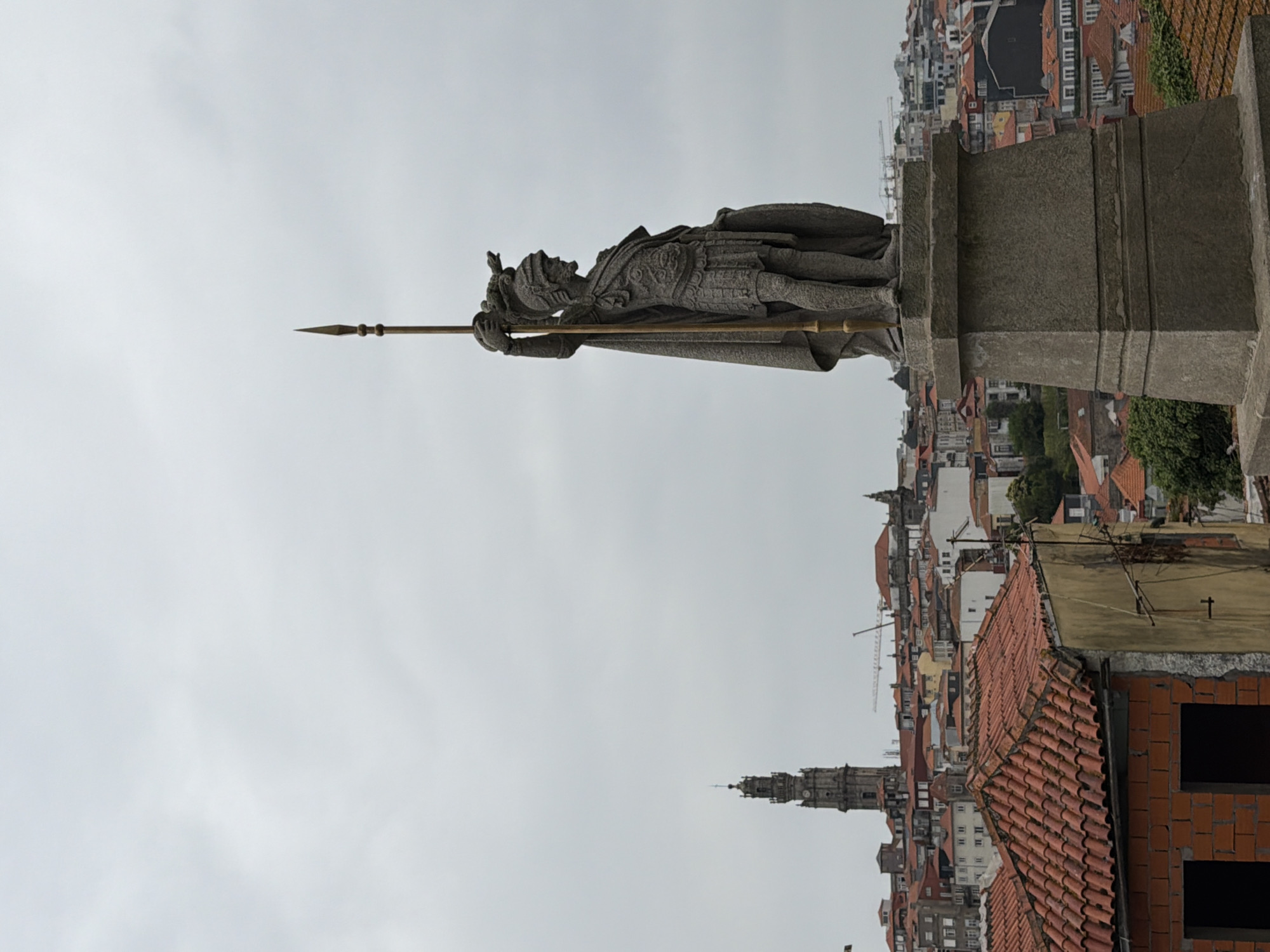 This media file captures a statue of a knight, likely a historical or symbolic figure, standing atop a stone pedestal. The knight is depicted in armor, holding a spear horizontally, suggesting a pose of readiness or vigilance. The background reveals a panoramic view of a city, characterized by a dense cluster of buildings with red-tiled roofs, indicative of an urban European setting. In the distance, a prominent clock tower rises above the cityscape, a common architectural feature in historic European cities. The sky is overcast and a light gray, suggesting a cloudy or perhaps misty day. The overall atmosphere appears to be one of solemnity and historical contemplation, with the statue serving as a focal point against the backdrop of the bustling city.
The context provided points to Porto, Portugal, as the location. This suggests the statue might be a monument within the city, possibly overlooking the Douro River or a significant square. The architecture visible, particularly the red-tiled roofs and the style of the clock tower, is consistent with the visual characteristics of Porto. No visible text is present in the image.
