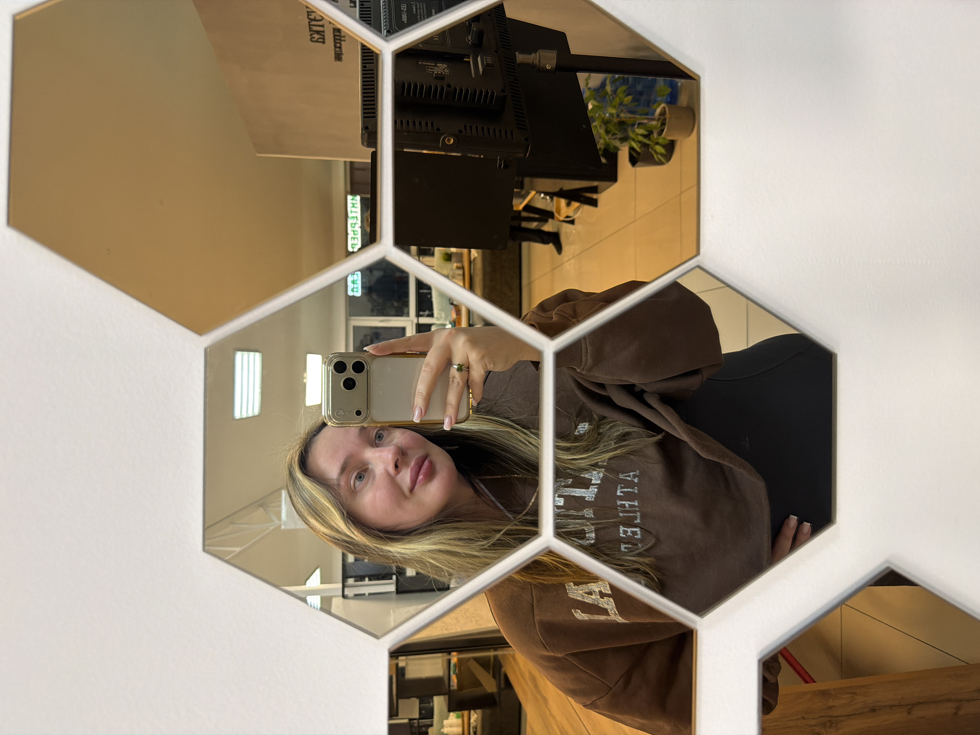 An image displays a mirror selfie of a young woman, captured within a decorative arrangement of hexagonal mirror panels on a wall. The photo is rotated 90 degrees counter-clockwise, showing the woman's head and torso reflected in the central panels. She is holding a smartphone, visible in the reflection, with her right hand, suggesting she is taking the picture.
The woman has long, highlighted blonde and light brown hair, fair skin, and light-colored eyes, with a neutral to slightly smiling expression. She is wearing a casual brown hoodie or sweatshirt. On her top, reflected in the mirror, is reversed text that appears to spell "ATLANTA". A ring is visible on her right ring finger.
The mirror assembly features several clear, reflective hexagonal panels alongside others that possess a warm, golden, or bronze tint, creating an aesthetic, fragmented view of the surroundings. Beyond the woman's reflection, the mirror panels show elements of an indoor setting, possibly in Chaikovsky, Russia. These reflections include electronic equipment, possibly a studio light or monitor, on a stand, a potted green plant, and hints of wooden surfaces. Other blurred and reversed text fragments like "SAJIKA" and "AHJIEPED" can be seen on background items within the reflections. The overall scene suggests a well-lit interior, possibly a modern living space, studio, or commercial establishment.
