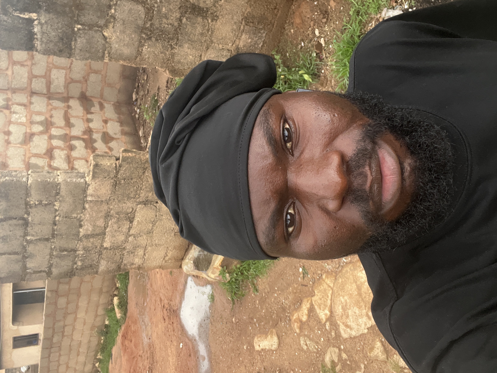 The image is a close-up, portrait-style photograph of a man with dark skin, a beard, and a black head covering. The man is looking directly at the camera. He is wearing a black shirt. Behind him is a wall constructed of concrete blocks and a dirt ground with patches of grass and a small puddle. The location is Gura Top, Nigeria. A window is partially visible on the lower left corner of the image.