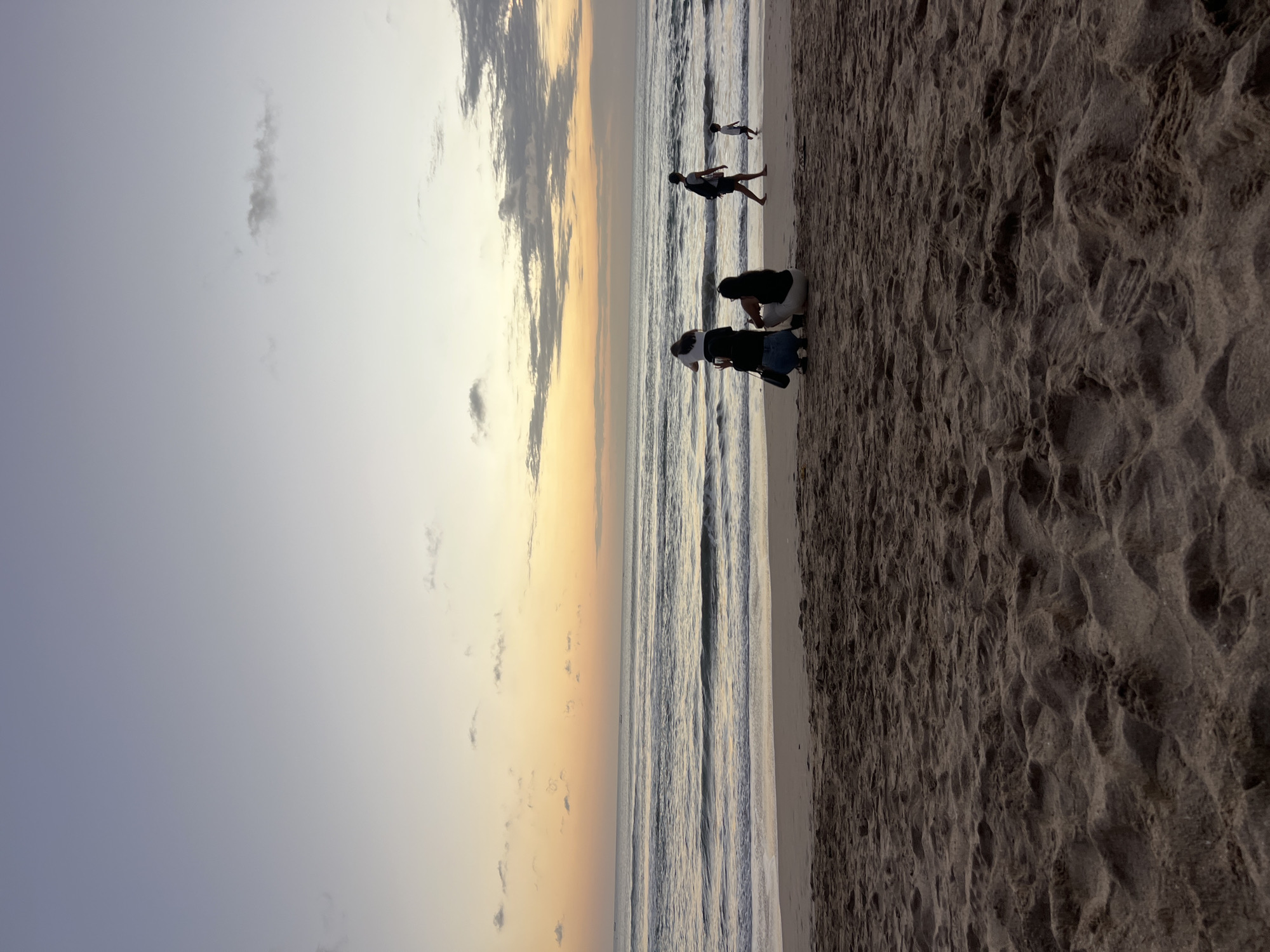 The image shows a tranquil beach scene at dusk in Kuta, Indonesia. The sky transitions from a pale blue at the top to warm orange and yellow hues near the horizon, indicating either sunrise or sunset. Wispy clouds are scattered across the sky. The sea exhibits gentle waves rolling onto a sandy beach. In the foreground, the sand is textured with footprints and ripples.
Several individuals are visible on the beach. Two people are sitting closer to the viewer, with one holding what appears to be a phone, possibly taking a picture. Further down the beach, towards the water, a person is walking away from the viewer, carrying a bag over their shoulder. A small child is also seen running near the water's edge. The overall atmosphere is peaceful and serene.