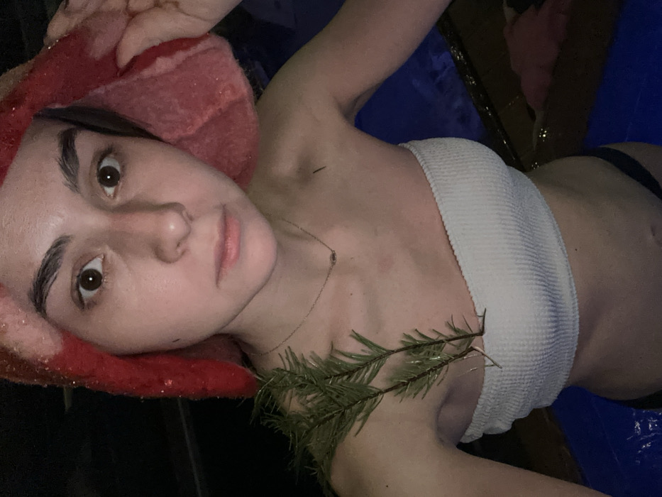 A female individual, appearing to be in her late teens or early twenties, is depicted in a medium close-up, partially submerged in water, presumably a pool or hot tub, at night or in a dimly lit indoor setting in Базарово, Russia. She is wearing a white, ribbed strapless bikini top and dark-colored bikini bottoms. Her head is wrapped in a textured, bright red fabric head covering, possibly sparkling or sequined. She gazes directly into the camera with wide-open eyes and a neutral expression. A thin, silver-colored necklace with a small, dark pendant is visible around her neck. Two green coniferous branches are positioned on her left shoulder and upper chest, tucked into the top of her bikini. A small, dark, linear object is visible on her right clavicle area. The water surface is blue, and the background is dark and indistinct.
