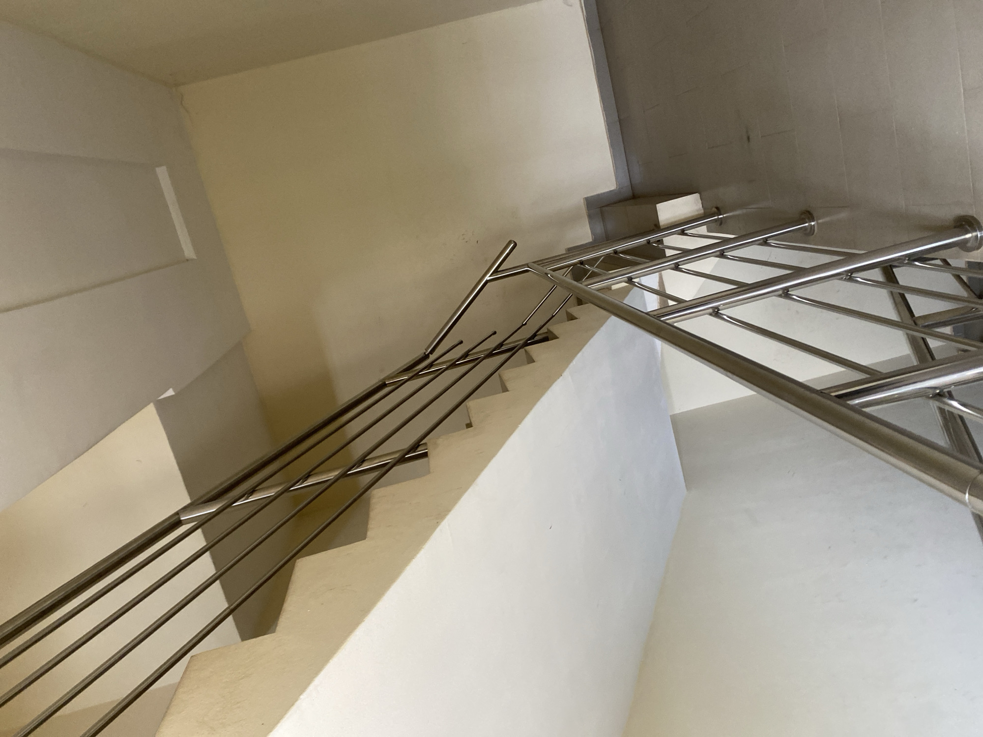 The image depicts an interior staircase with white stairs and walls. A metal handrail runs along the stairs. The scene appears to be in an institutional setting, possibly a public building. The location is undetermined.