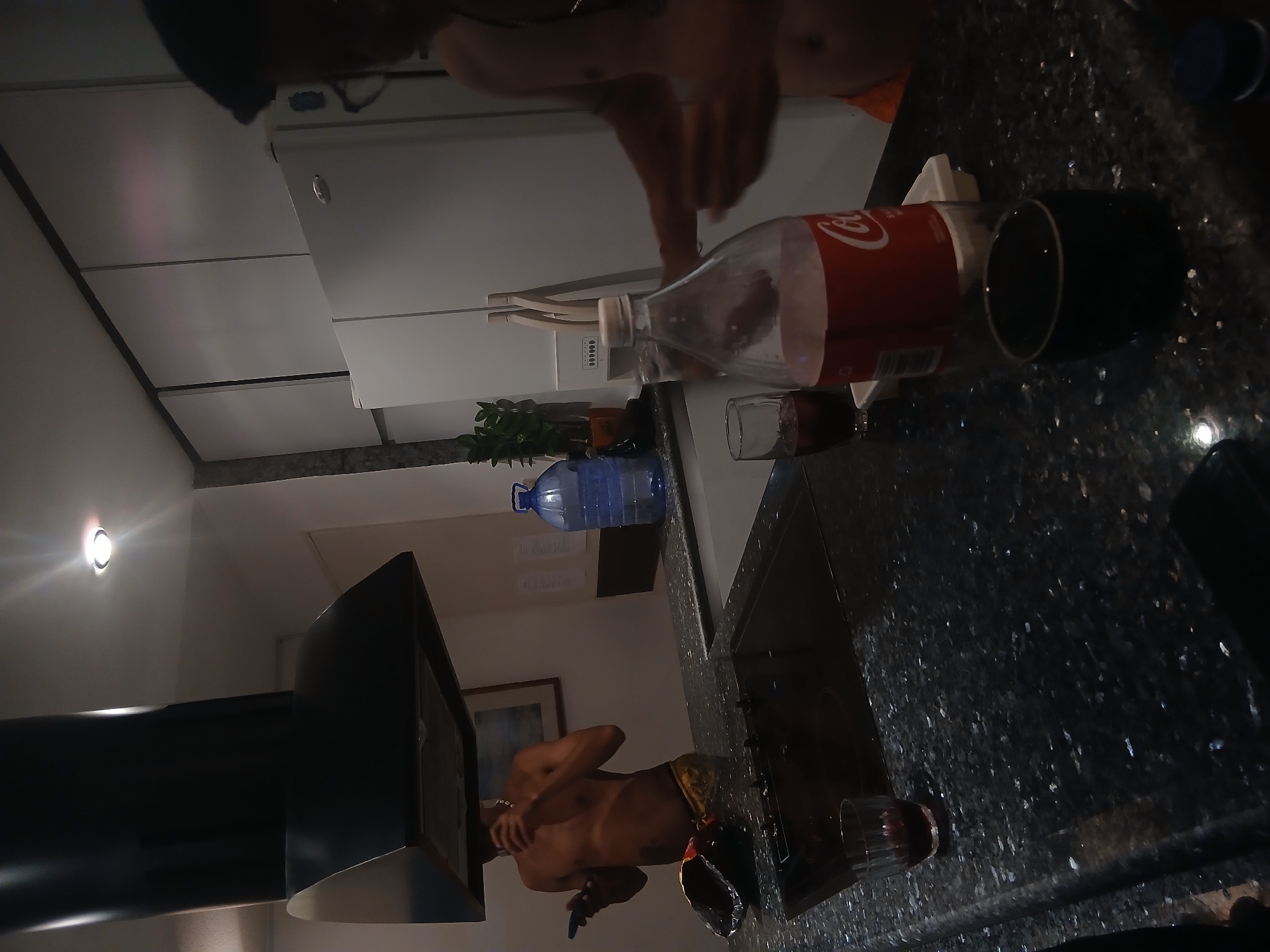 The image depicts an indoor scene, possibly in Porlamar, Venezuela. A kitchen countertop is visible, cluttered with a Coca-Cola bottle, a black mug, and a glass. A partially unclothed individual stands near a kitchen sink with a window. The person is shirtless. A bottle of water and some leafy plants are visible further back on the counter. A range hood extends out above the standing person.