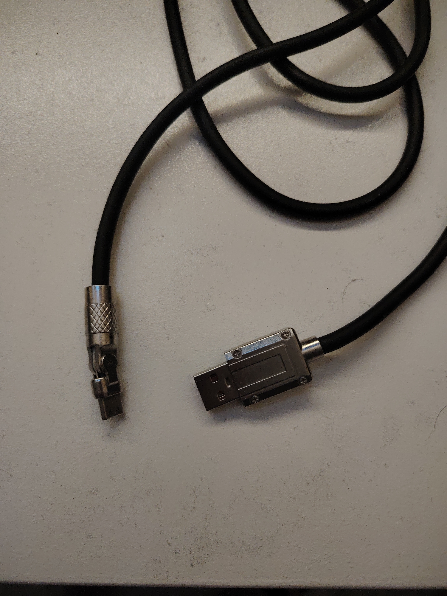 A black insulated electrical cable with two distinct metallic connectors is positioned on a light-colored, subtly textured surface. One end of the cable terminates in a silver-toned, knurled cylindrical collar, from which a smaller, dark metallic male connector component extends, appearing either broken or modularly detached from a larger assembly. The other end of the cable is equipped with a standard USB Type-A male connector, housed within a rectangular, silver-toned metallic casing secured by visible Phillips head screws. The cable is loosely coiled across the surface, which exhibits minor particulate matter and subtle surface variations under diffuse overhead lighting. This scene is observed in Huntingdonshire, United Kingdom.