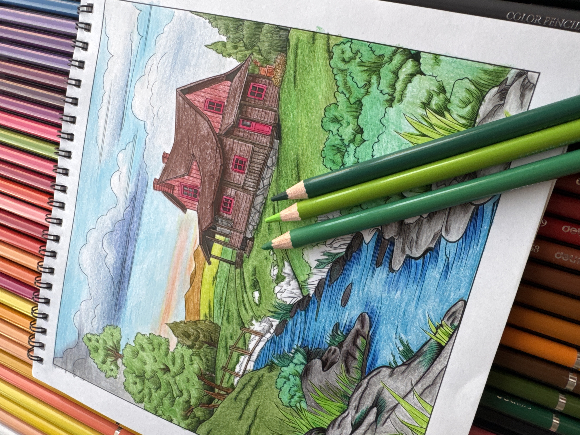 The image displays a colored pencil drawing of a rural landscape featuring a rustic wooden house with pink window frames and a red door, situated on a grassy hillside. A stream with blue water flows through the foreground, bordered by rocks and lush green vegetation. A wooden bridge crosses the stream in the mid-ground. The drawing is presented on a sketchbook page, with several green colored pencils lying on top of it. The sketchbook is placed on a surface covered with a large assortment of colored pencils, arranged in rows. The overall scene is brightly lit, suggesting an outdoor setting. There are no people or animals depicted in the drawing. The image does not provide any specific information about the city of Nha Trang Ward, Vietnam, as the scene is a generic landscape.