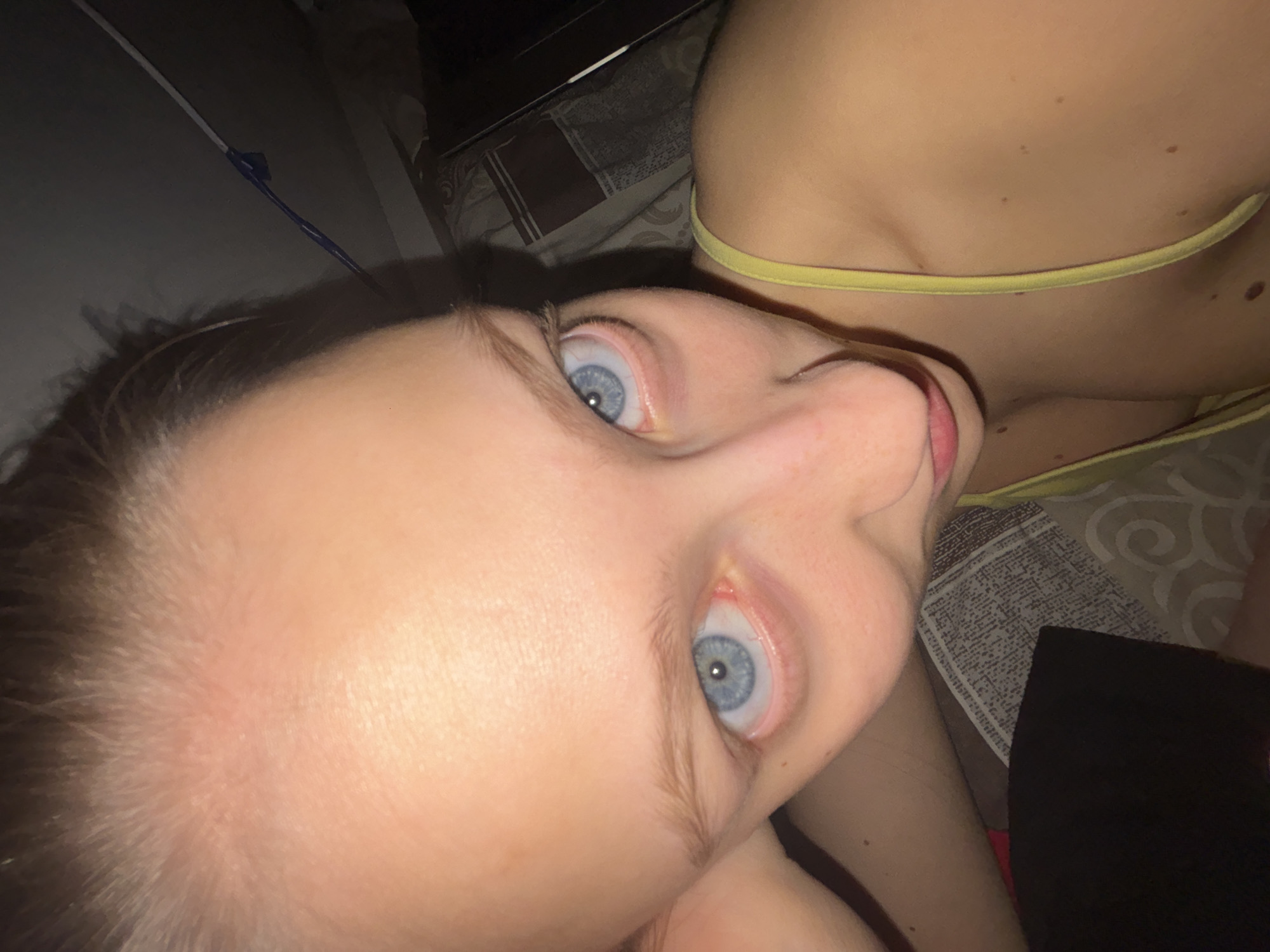 A close-up, angled view of a young woman's face and upper body. The woman has fair skin, light hair, and striking blue eyes. She is lying on her side, with her head tilted upward towards the camera. She is wearing a yellow top with thin straps. The background consists of bedding with various patterns and colors, and a black object partially visible on the right side. The image does not contain any information that can reveal the location in the city of Perm, Russia. The image contains only essential details about people, subjects, objects, actions, interactions, and the environment.