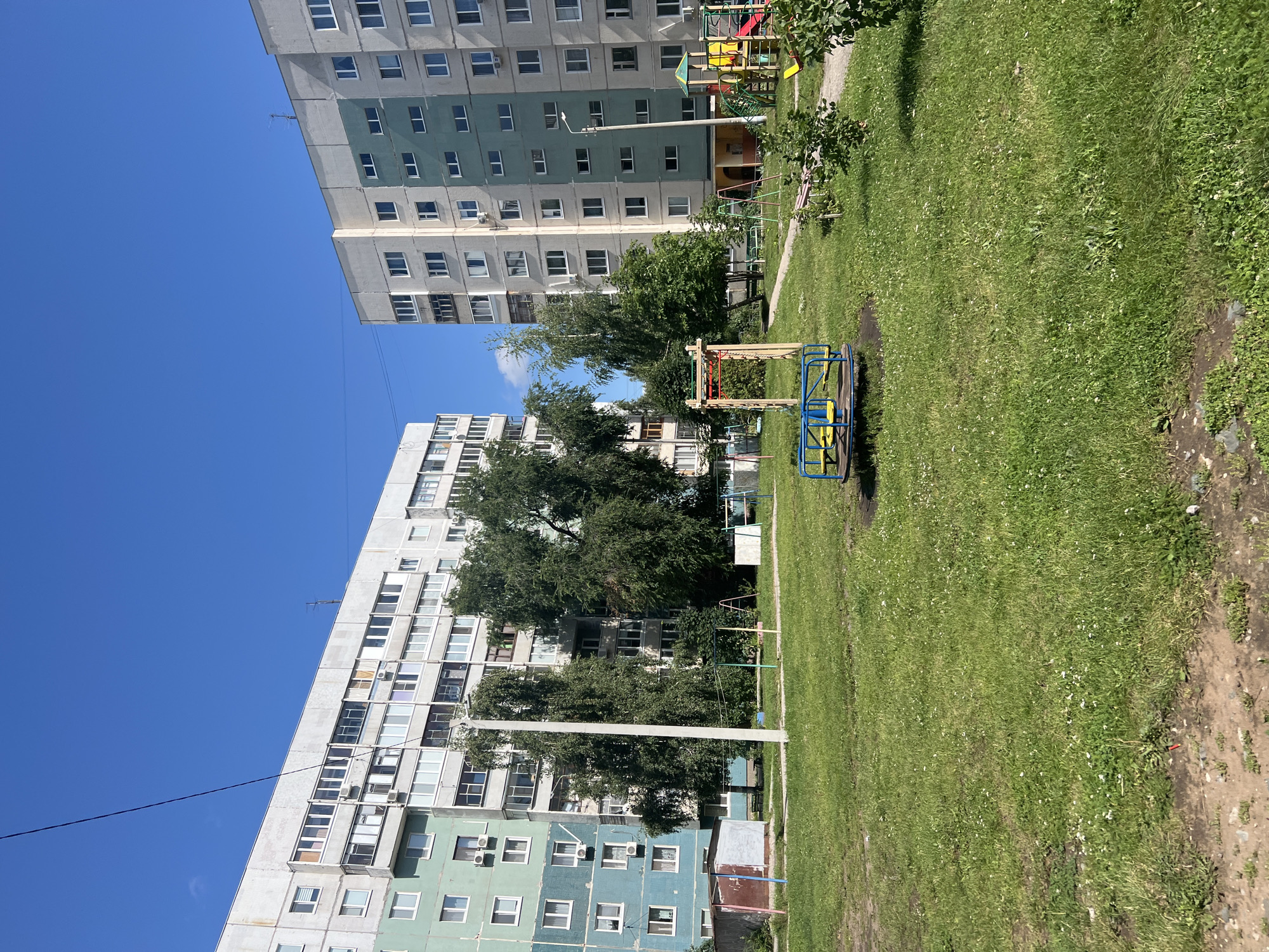 The image depicts a residential courtyard in Ulyanovsk, Russia, under a clear blue sky. Two multi-story apartment buildings, typical of Soviet-era construction, dominate the scene. The building on the left is primarily gray and white with some sections painted in a teal-blue color, while the building on the right is mostly gray and white with turquoise accents. Both buildings feature numerous windows and balconies.
In the foreground, a grassy area with scattered small white flowers covers the ground. A children's playground is situated in the courtyard. A blue and yellow merry-go-round is prominently positioned in the center of the frame. To the right of the merry-go-round, a larger playground structure with red, yellow, and green elements is visible, along with swings. Mature trees with lush green foliage are interspersed throughout the courtyard, providing shade and breaking up the expanse of grass and buildings. A dirt path winds through the playground area. In the background, near the left building, a small, utilitarian shed with a red and gray exterior is present. A utility pole stands near the center of the image. No people are visible in the scene. The overall impression is a somewhat dated but functional urban residential environment.