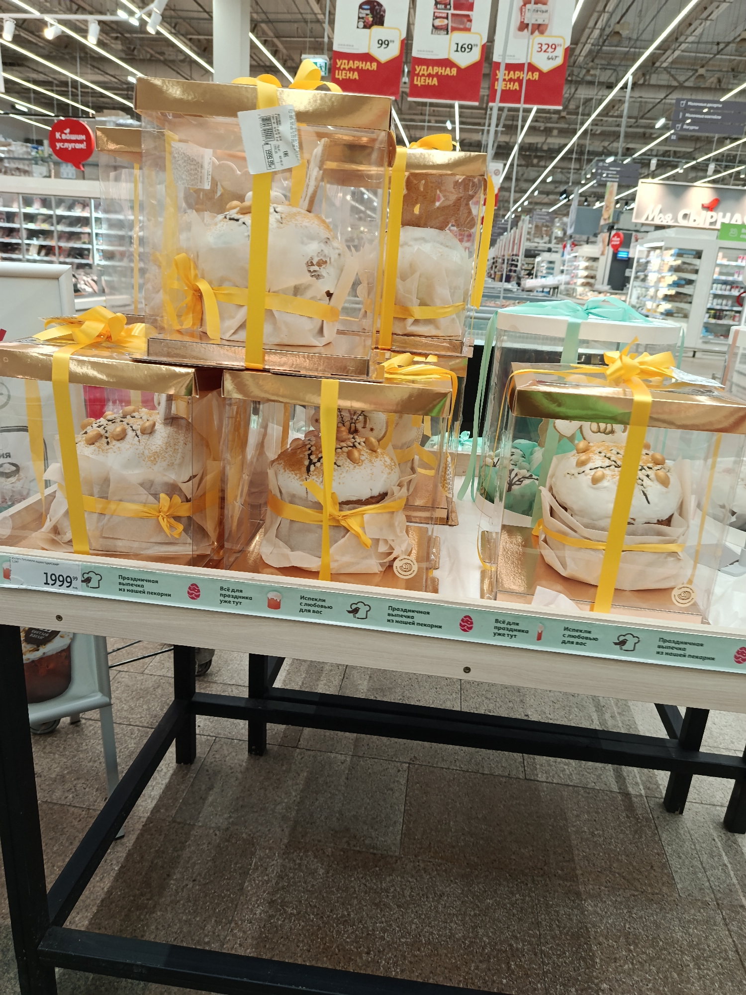 The image captures a supermarket display featuring festive baked goods, likely traditional Russian Easter cakes (Kulich). Multiple cakes are individually packaged in clear plastic boxes with gold tops and yellow ribbons, arranged on a wooden stand. Some boxes feature light blue/mint green ribbons. The cakes themselves are topped with white icing and various nuts or sprinkles.
In the background, typical supermarket aisles are visible with shelves stocked with products and bright overhead lighting. Several red signs hang from the ceiling, displaying "УДАРНАЯ ЦЕНА" (Bargain Price) along with prices such as "99", "169", and "329". A red speech bubble sign reads "К вашим услугам!" ("At your service!"). Further down the aisle, a sign partially reads "Моя СЫРНАЯ" (My CHEESE).
The front edge of the display stand contains promotional text and icons. A price of "199.99 РУБ." is visible, alongside phrases in Russian: "Праздничная выпечка из нашей пекарни" (Festive pastry from our bakery), "Всё для праздника уже тут" (Everything for the holiday is already here), and "Испекли с любовью для вас" (Baked with love for you). These texts, along with small Easter egg and baker's hat icons, clearly indicate a seasonal Easter promotion from the store's in-house bakery. The scene depicts a retail setting in Moscow, Russia, offering specialty items for a holiday.