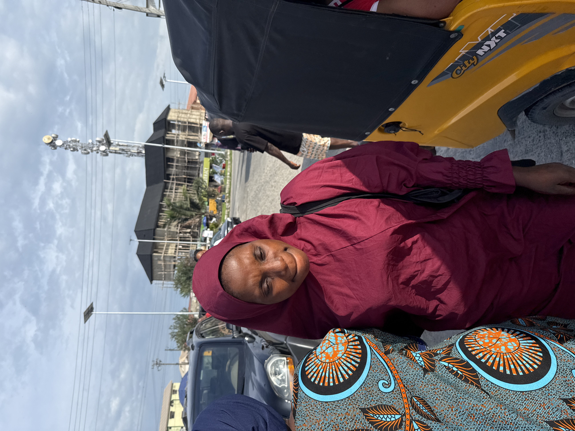 The image shows a woman wearing a maroon hijab and dress, seated in what appears to be a yellow "City NXT" tricycle taxi, possibly in Ado, Nigeria. She is smiling and looking towards the camera. Beside her is a person wearing blue and orange patterned clothing. In the background, a building under construction is visible, along with a communications tower and other vehicles. The weather appears overcast.