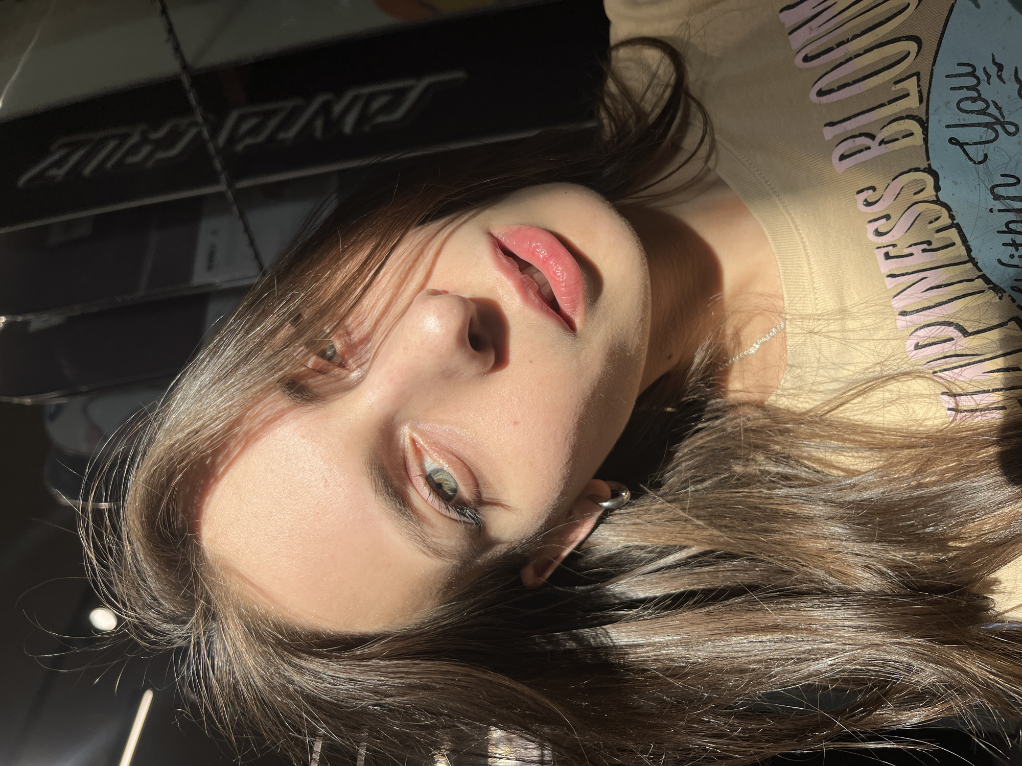 A portrait of a young woman with light brown hair is angled towards the camera. Her skin is fair and her lips are slightly parted and pink. She has green eyes and is wearing a small hoop earring. She is wearing a light-colored t-shirt with text, partially obscured by the angle, but visible enough to read "princess blossom". Sunlight illuminates parts of her face and hair. Dark, reflective surfaces are visible in the background. The location in the city of Krasnoyarsk, Russia, is not deducible from the image.