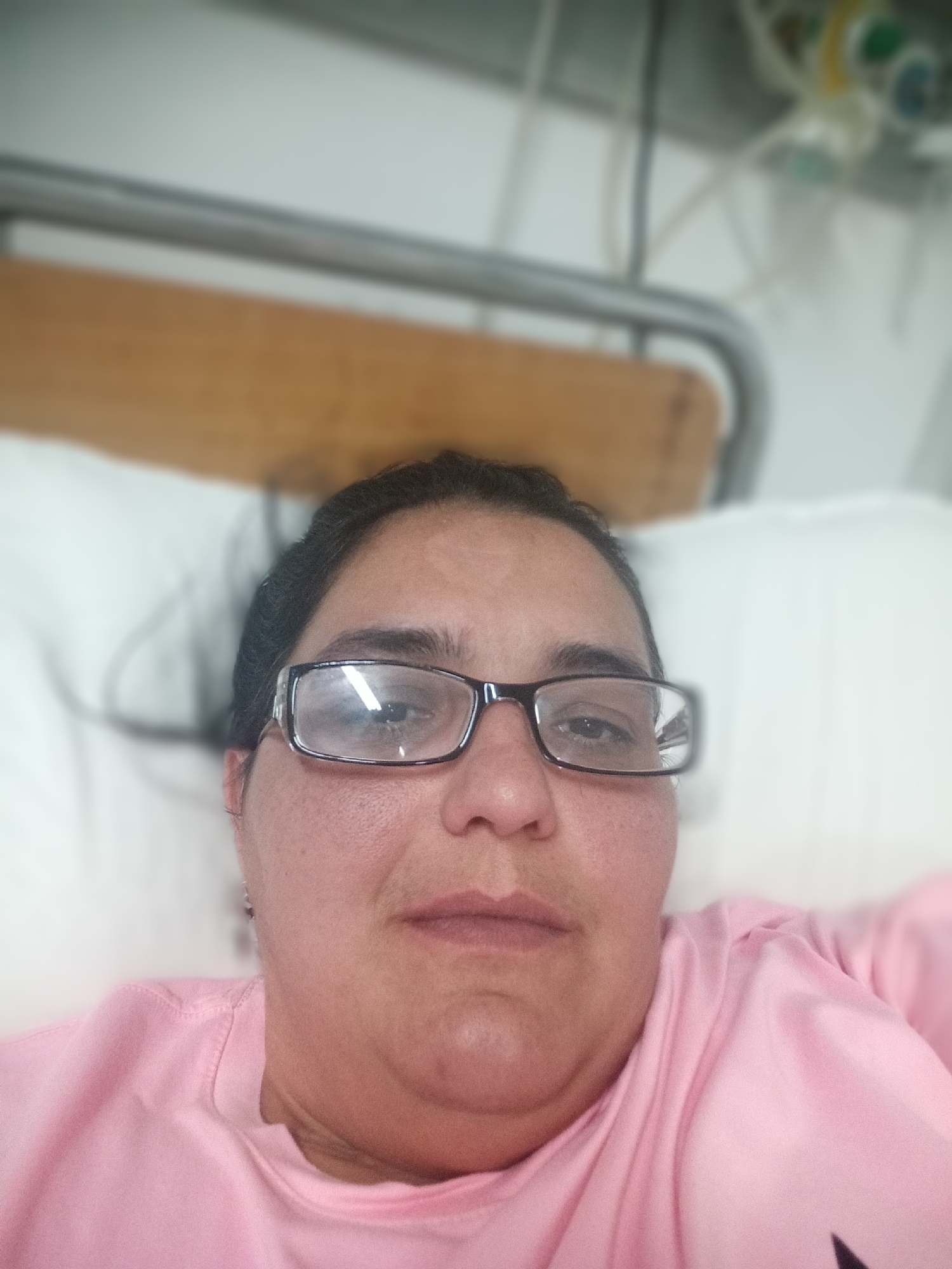 A close-up, selfie-style photograph captures a woman from the chest up, lying in a hospital bed. She is wearing a pink crew-neck shirt and glasses with dark frames. Her dark hair is pulled back, and her expression is neutral. The background shows a white pillow, a wooden headboard, and medical equipment hanging above, suggesting a hospital room. The overall scene is underexposed and out of focus, with the woman's face being the sharpest element. The exact location in Pleven, Bulgaria, cannot be determined from the image alone.