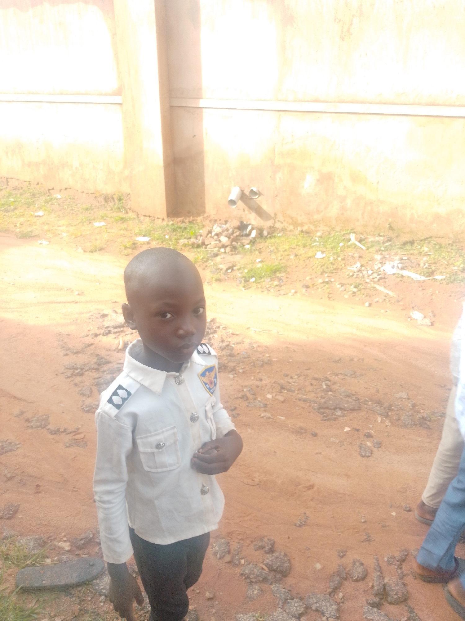 A medium shot captures a young boy standing in a dirt area next to a light-colored wall. The boy is wearing a white shirt with epaulets and a patch on the left chest, and black pants. His hands are visible, with one resting on his hip and the other in front of his body. In the background, the wall has a white horizontal pipe near the top and some smaller pipes or conduits lower down. The ground is composed of reddish-brown dirt with scattered stones and some green grass. To the right of the boy, the lower legs and feet of two other individuals are visible, one wearing light-colored pants and brown shoes, and the other wearing blue pants and brown shoes. The lighting suggests it is daytime.