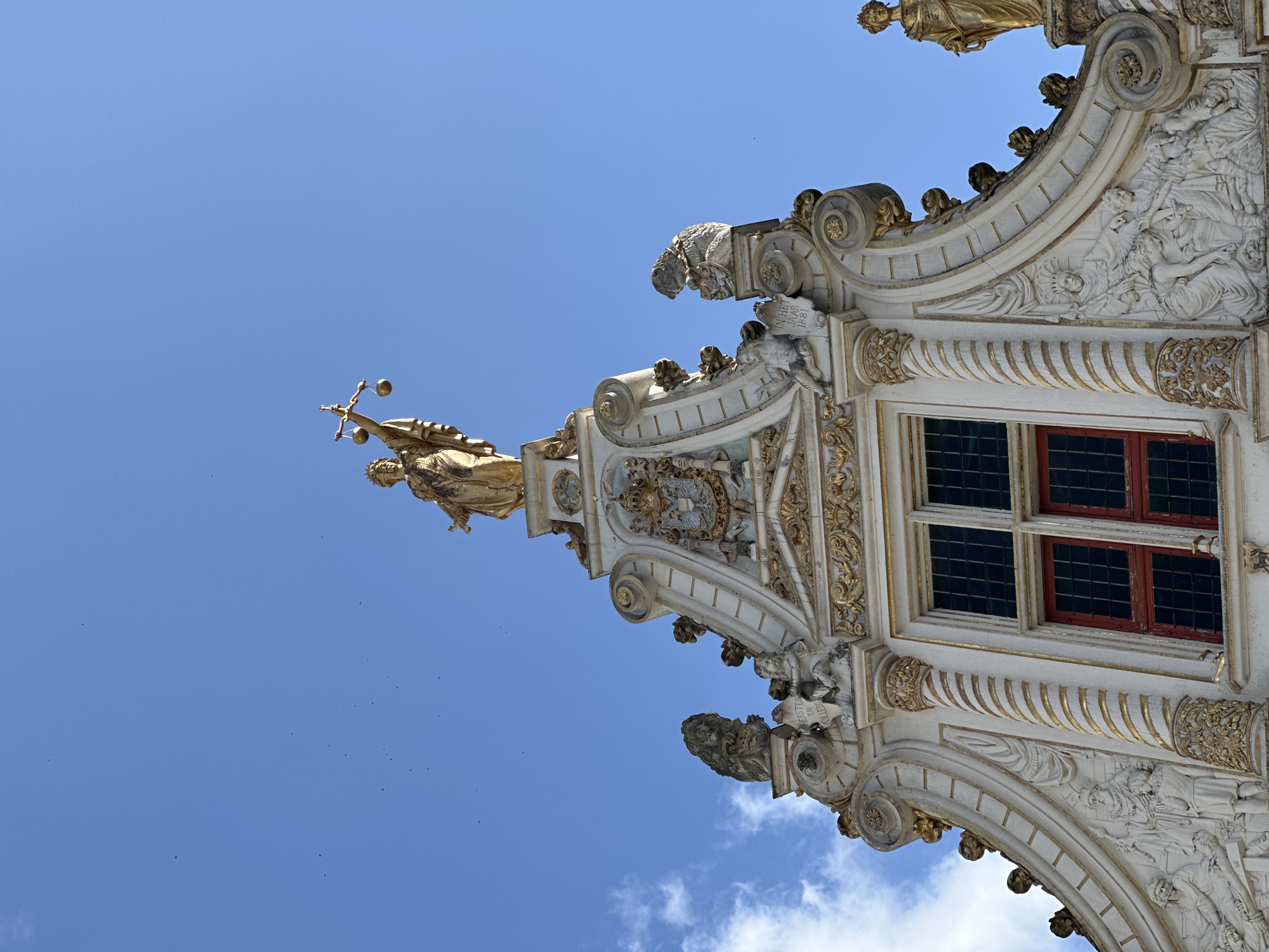 The image shows the detailed facade of a building in Bruges, Belgium, under a clear blue sky with scattered clouds. The architecture is ornate, with a triangular pediment topped by a golden statue of Justice holding scales. Below this statue is a coat of arms and further decorative elements, including reliefs of figures and lions. A large window with red frames is visible on the facade. The building's exterior is adorned with intricate white stone carvings and gilded embellishments. The photo is taken from a low angle, looking up at the building.