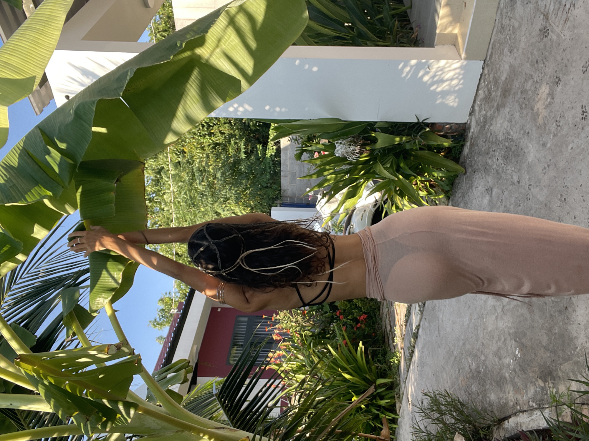 A woman stands outdoors in a garden setting, with large banana leaves framing the top portion of the image. She is viewed from behind, wearing a dark criss-cross back bralette and a sheer, flowing beige skirt. Her arms are raised, with her hands reaching up to touch the banana leaves. The background shows lush green foliage, some red flowers, a red-roofed building, and a white pillar. The ground is a concrete pathway. The lighting suggests it is daytime with clear skies. The specific location in Mũi Né, Vietnam is not identifiable from the visual information alone.