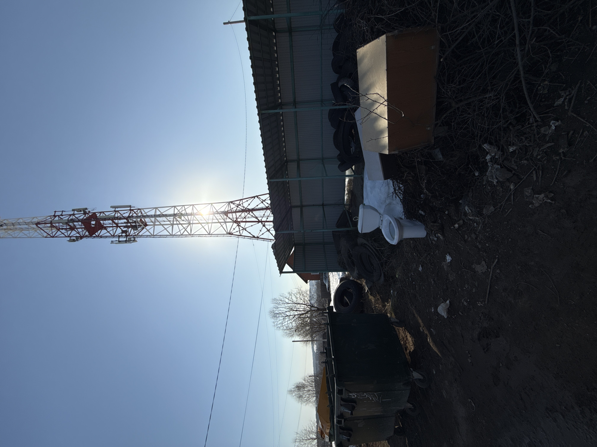 The image shows an industrial site in Votkinsk, Russia. A red and white communications tower stands next to a metal structure, possibly a storage shed, filled with tires. An old wooden box and a white toilet sit discarded on the ground next to the shed. A large, dark container, likely a dumpster, is located near the bottom of the frame. The ground is bare and appears to be a dirt or gravel surface. The sky is clear blue with the sun shining through the communications tower. Bare trees are visible in the background. There are no people visible in the frame.