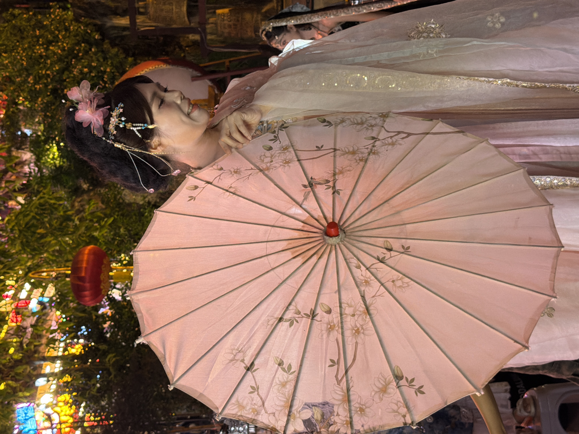 In Yuzhong District, China, a young woman adorned in traditional Chinese attire, possibly a Hanfu, is featured prominently in the foreground, holding a delicate, light pink umbrella. The umbrella is open and displays an intricate floral pattern in lighter shades of pink and beige. She is wearing a light-colored outer robe with sheer sleeves, embellished with golden sequins and floral embroidery, layered over a pink garment. Her hair is styled in an updo, decorated with a pink flower and delicate bejeweled hairpins. She is smiling and looking downwards, her expression gentle. Behind her, a blurred figure of another woman, also in traditional attire with a dark veil and intricate headwear, is visible, suggesting a festive or cultural event. The background is illuminated by a multitude of colorful lights, including red lanterns and multicolored string lights, creating a vibrant and festive atmosphere. The overall scene captures a moment of cultural immersion and celebration in a dynamic urban environment.