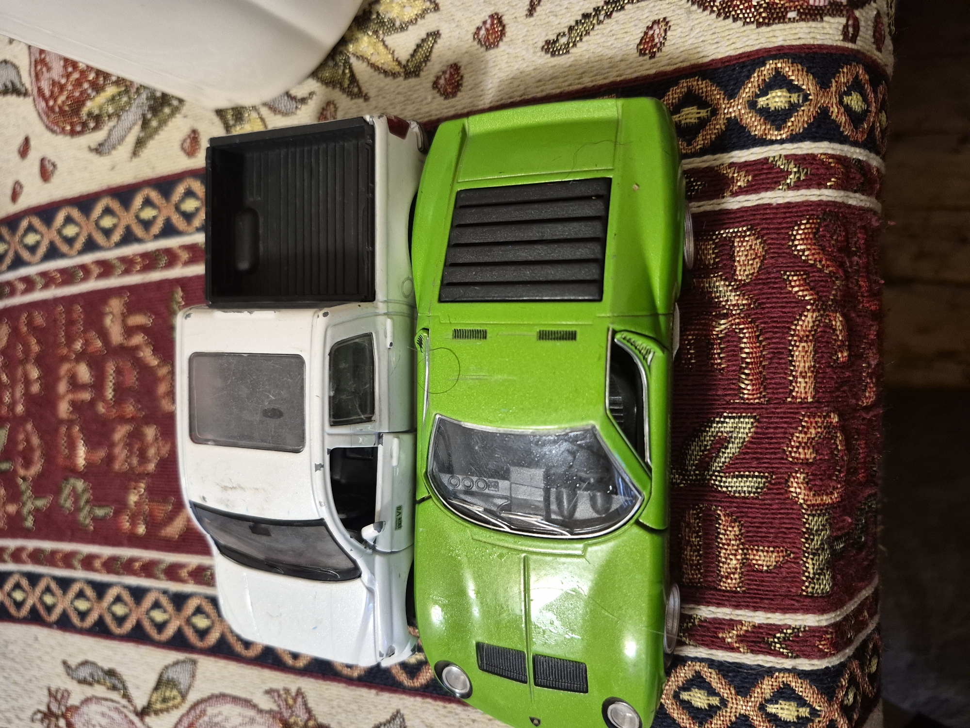 An overhead view captures two well-used toy cars resting on a richly patterned fabric, likely a rug or tapestry, with a plain white surface visible in the background. The scene depicts a still life indoors, potentially in Yerevan, Armenia, given the decorative style of the fabric.
To the left, a white pickup truck model is visible. Its open truck bed, featuring a textured black liner, is prominent. The side of the cab shows a window and an open door or a broken part, revealing interior details. On its side, a small silver emblem clearly displays the text "V8". The truck appears to have been played with, showing some scuffs and wear.
To the right, a lime green sports car, resembling a classic Lamborghini Miura, lies alongside the truck. It features distinctive horizontal louvers over what would be the engine compartment at the rear, and round headlights on the front. The windshield provides a glimpse of the dashboard, which includes three circular gauges. This car also exhibits signs of wear, with scratches and small paint chips across its metallic green finish.
The patterned fabric beneath the cars features intricate designs in shades of deep red, gold, black, and cream, with geometric and stylized motifs. This traditional pattern adds a cultural context to the otherwise simple arrangement of toys. No activity is taking place; the cars are static. The lighting suggests an indoor setting, likely under artificial light.