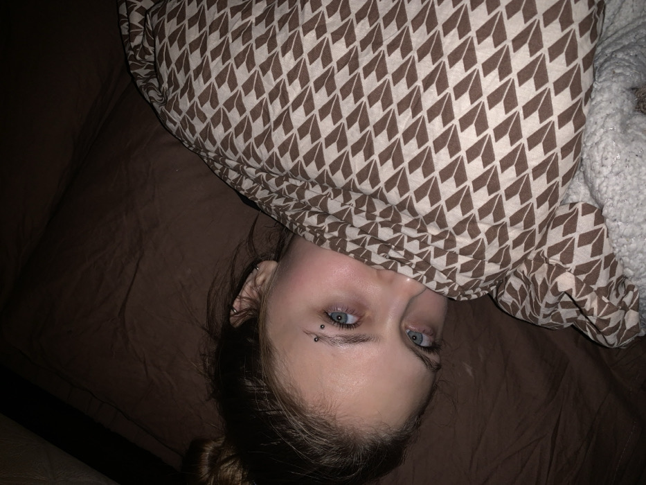 The image depicts a close-up, dimly lit scene within an indoor setting in the city of Фоки, Russia. A single individual, appearing to be a young female, is lying down and looking directly into the camera. Her face, primarily her eyes and forehead, is visible, with the lower portion obscured by bedding. She has light blue eyes and an eyebrow piercing consisting of two silver-colored studs on her left eyebrow. Her hair, pulled back, is visible on the upper left. She is partially covered by a patterned blanket featuring light brown, angled, geometric shapes on a darker brown background. A plain brown sheet or blanket is visible behind her head and to the upper left. A white, textured material, possibly a fleece blanket, is visible at the bottom right of the frame. The lighting is low, suggesting an evening or nighttime setting.