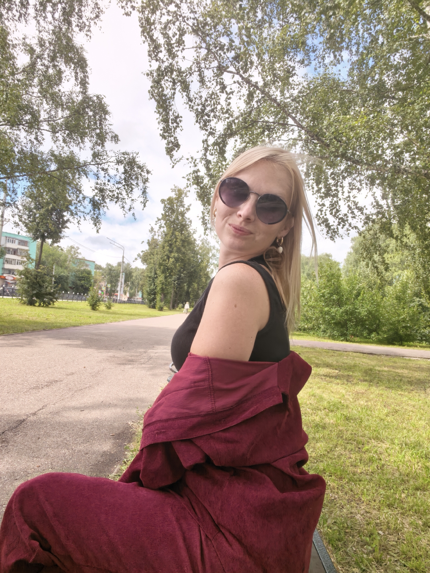 A blonde woman with sunglasses and large earrings is sitting outdoors, possibly on a bench, in a park-like setting with trees and a paved path. She is wearing a black top and a maroon outfit draped over her shoulders and lap. In the background, there is a street with buildings and more trees. The sky is visible through the branches of the trees. The image was taken in Sterlitamak, Russia.