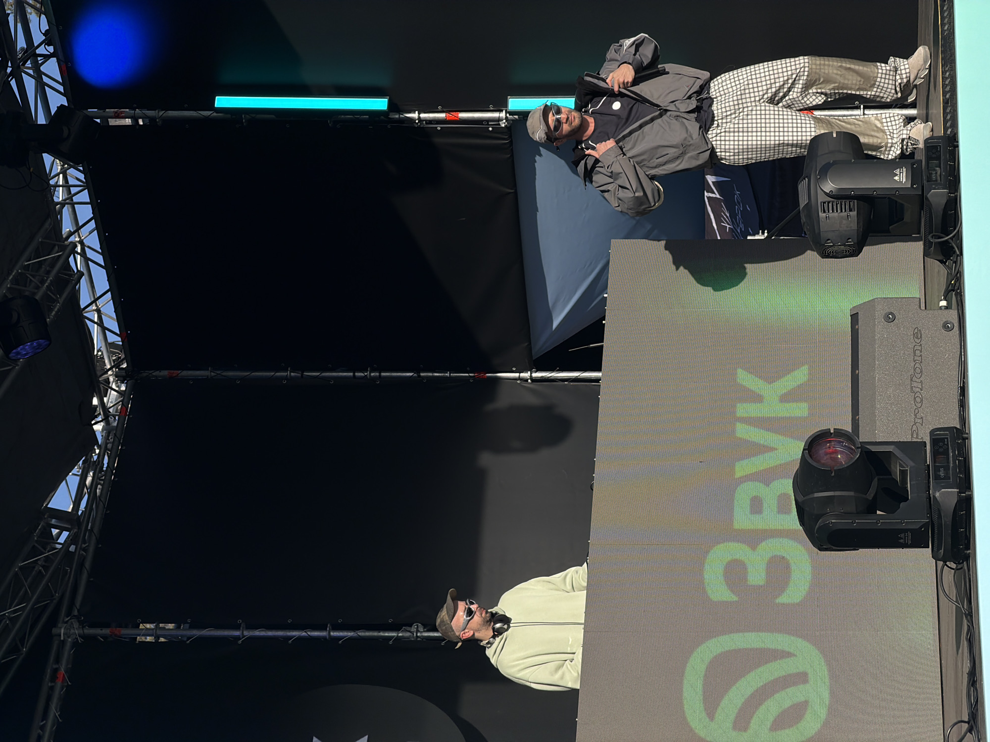 The image shows two men performing on a stage. The location is likely Озёрное, Russia, given the "Звук" logo on the screen, which is a Russian word meaning "sound." One man is wearing a light-colored jacket and headphones, while the other is wearing a grey jacket and checkered pants. They are both wearing sunglasses and holding microphones. The stage has lighting equipment and a backdrop with the "Звук" logo.