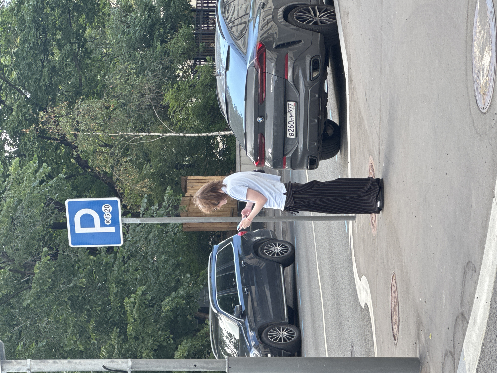 A person is standing on a sidewalk, looking at their phone. They are wearing a light blue t-shirt and black striped pants. Two cars are parked on the street behind them, a dark blue SUV and a dark gray BMW coupe. A blue parking sign with a white "P" and numbers "10 15 20" is visible in the background. The scene is set in a city, with trees lining the street and buildings visible in the distance. The license plate of the BMW is B260HM977, indicating it is registered in Moscow, Russia. The overall impression is that of an everyday urban scene.