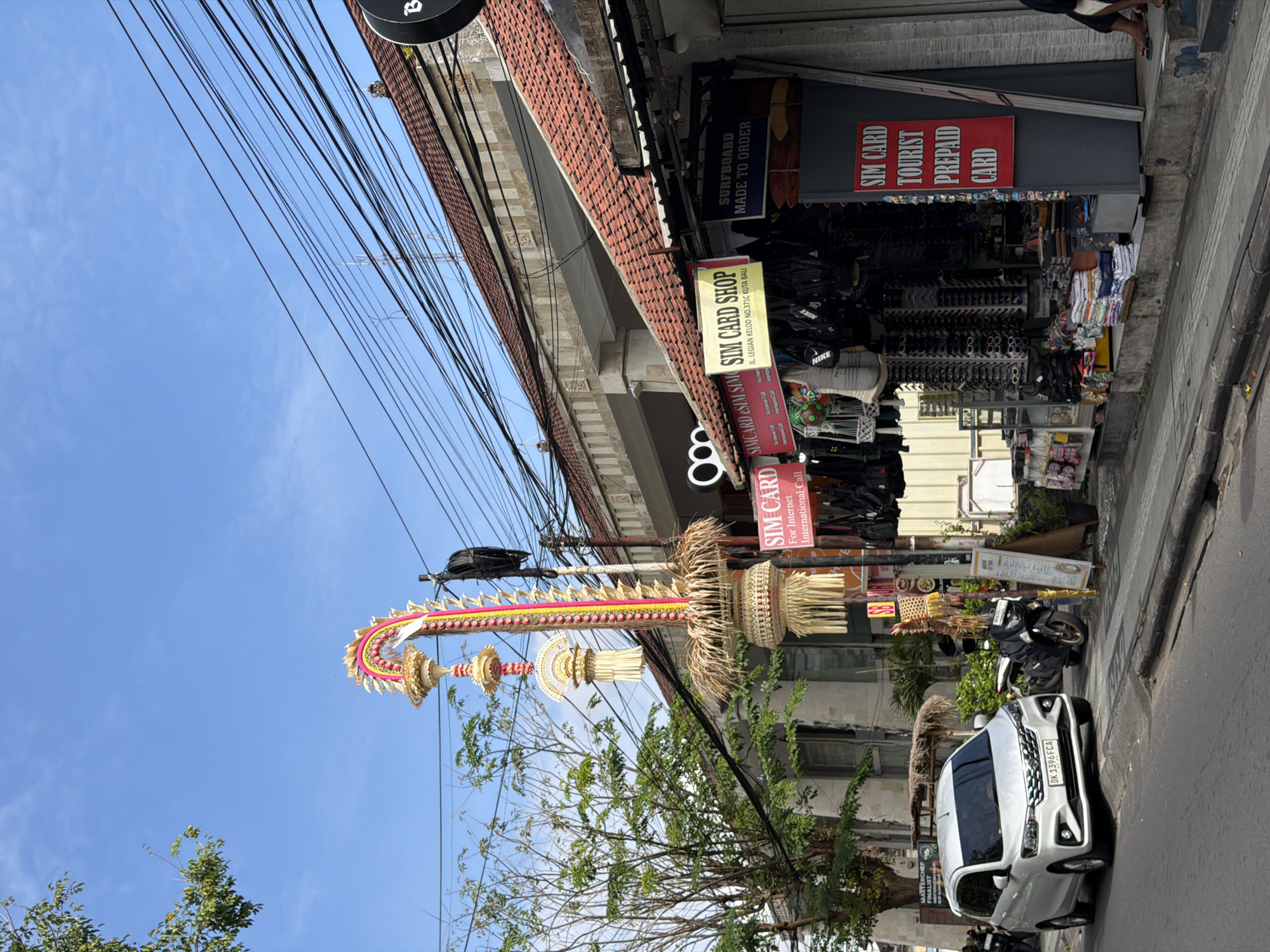 In Kuta, Indonesia, a street scene unfolds under a clear blue sky. A white minivan with the license plate DK 1396 FCA is parked on the right side of the road. Beside it, a black motorcycle is also parked. In front of a building with a red-tiled roof, a SIM card shop is visible, with various items like sunglasses and bags displayed for sale. A tall, decorative structure with intricate weaving and colorful accents stands near the shop, likely a traditional Balinese ornament. Overhead, a tangle of electrical wires crisscrosses the sky. The scene captures a typical day on a busy street in Kuta, showcasing a mix of transportation, commerce, and local culture.