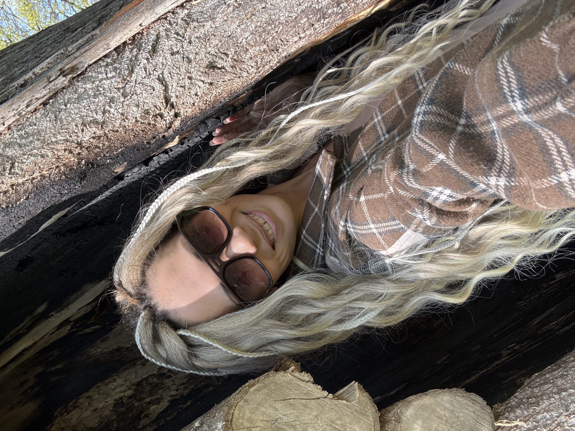 A woman wearing sunglasses and a brown and white plaid shirt is lying inside a partially burned-out log. The interior of the log is charred black. Her blonde hair, styled with white rope-like braids, spills out around her. The photograph appears to have been taken from within the log, looking up towards the sky, as some greenery can be seen. The bottom of the log where her head lies contains several small, unburned logs. The location is Rusanovo, Russia.