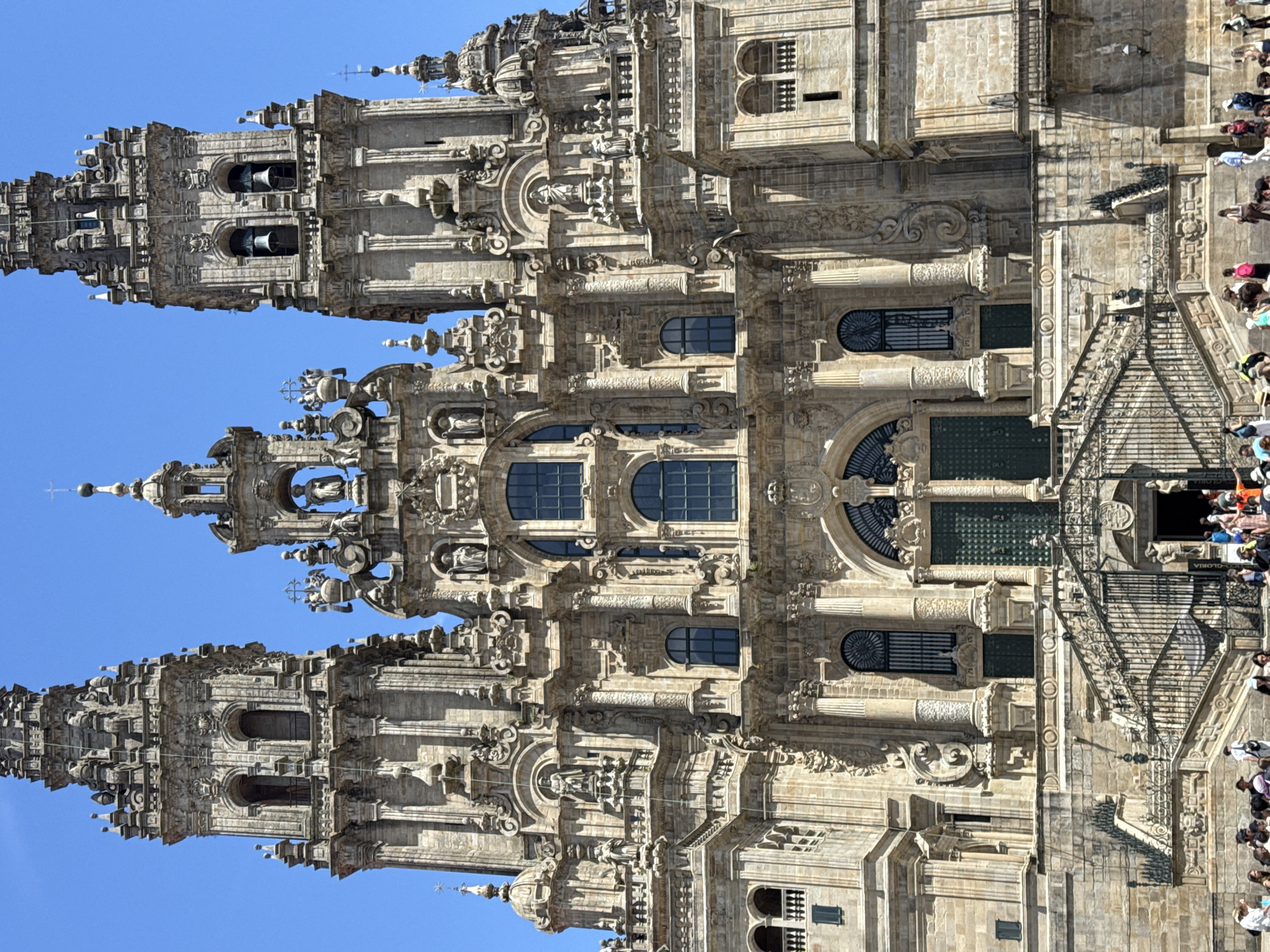 This is a vertically oriented photograph of the facade of the Santiago de Compostela Cathedral in Spain. The architecture is highly ornate, featuring intricate stone carvings, columns, arched windows, and spires. Multiple bell towers are visible, with bells hanging in some of them. The stone is a light beige color, and the building is illuminated by bright sunlight, suggesting a clear day.
A group of people are gathered on and around a set of stairs leading up to one of the entrances. They appear to be tourists, some looking at the cathedral, others walking. Their attire suggests a warm day. The clear blue sky is visible behind the upper portions of the cathedral.
The scene appears to be a typical day in a popular tourist destination, with visitors exploring and admiring the historic cathedral. The time of day is likely midday or early afternoon, given the intensity of the sunlight and the long shadows cast on the building and ground.
There is some visible text on a plaque or sign near the entrance where the people are gathered, which reads "GLORIA". This likely refers to the "Puerta de la Gloria" (Portico of Glory), a famous entrance to the cathedral.
