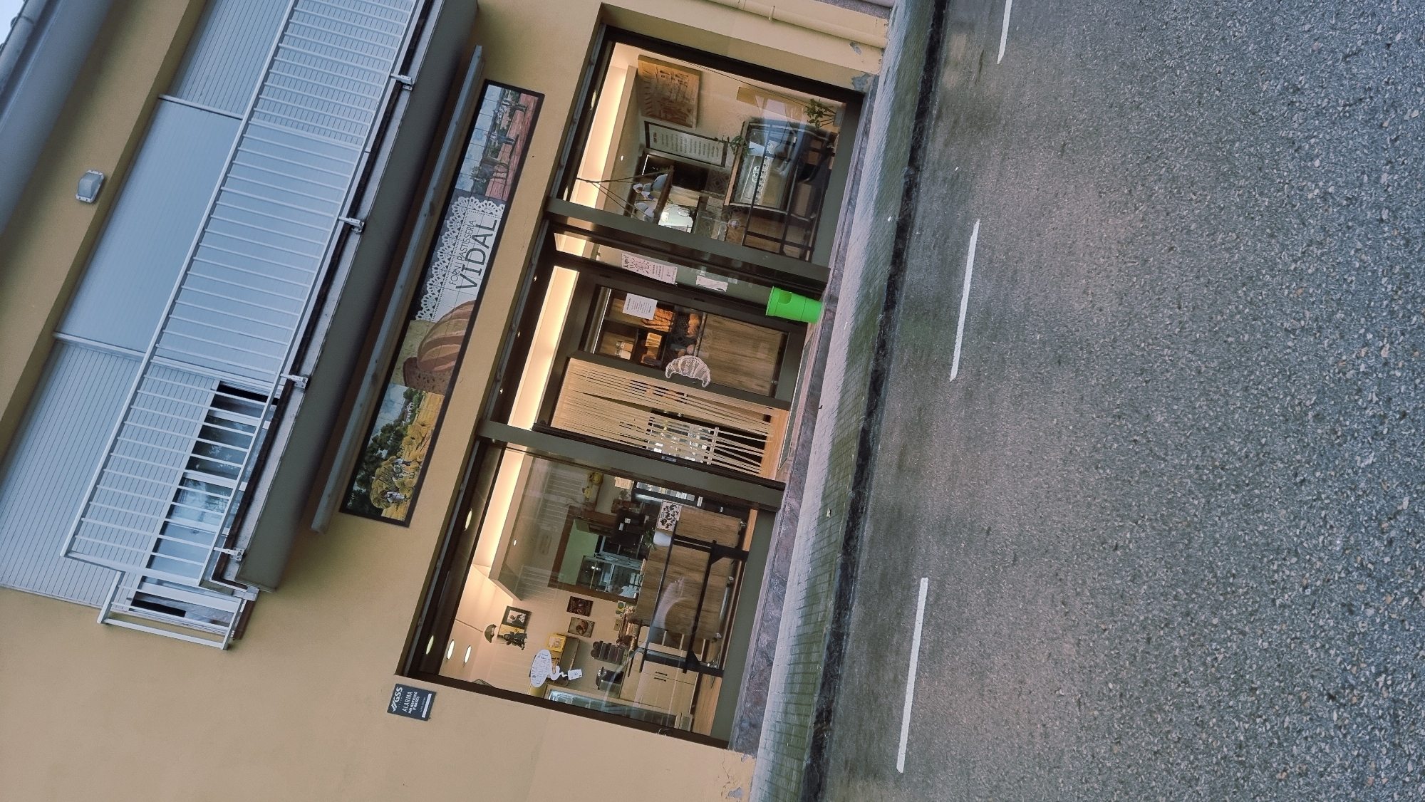The image shows the exterior of a bakery and pastry shop named "VIDAL FORN I PASTISSERIA" in Sant Joan Les Fonts, Spain. The shop is on the ground floor of a building with a yellow facade and a balcony above. The entrance features large glass windows and doors, revealing the interior of the shop, which appears to be well-lit and stocked with baked goods. A green trash can is visible outside the entrance. The street in front of the shop is wet, suggesting recent rain, and has white lane markings. A sign indicating an alarm system is mounted on the building near the entrance. The overall scene depicts a commercial establishment in a European town.