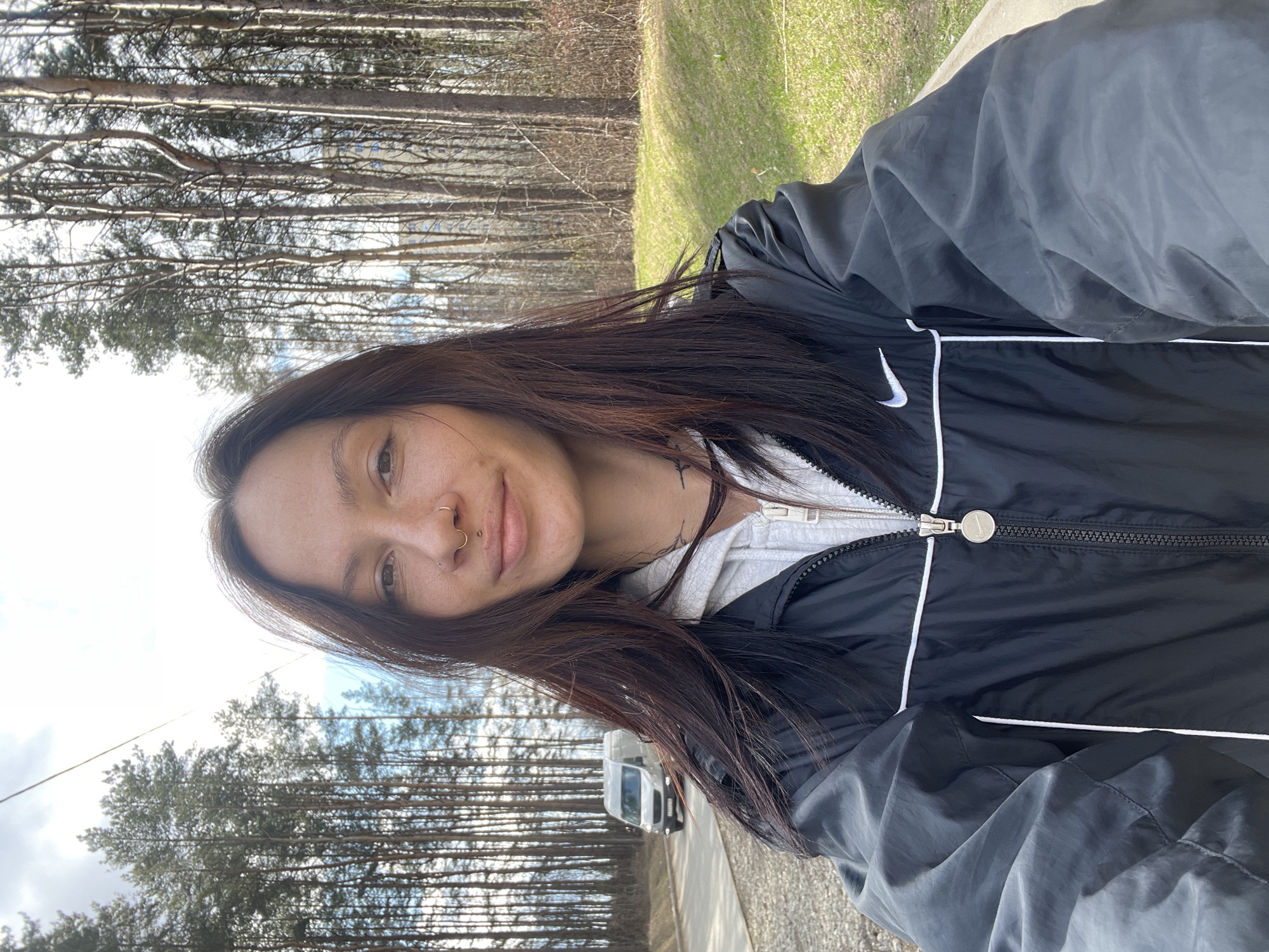 A selfie captures a woman outdoors in Izhevsk, Russia, under bright, partly cloudy skies. She is positioned in the foreground, smiling gently and looking directly at the camera. Her long, dark brown hair cascades over her shoulders. She wears a black Nike jacket with a white swoosh logo on the chest and white piping along the seams, layered over a grey hooded top. Visible accessories include a septum ring and a nostril ring.
The background features a dense stand of tall, slender trees, possibly conifers, suggesting a wooded area or forest. To her right, a patch of vibrant green grass is visible, hinting at a natural or park-like setting. A paved road or path runs through the scene, with a white van partially visible in the distance among the trees. The time of day appears to be daytime, with ample natural light.