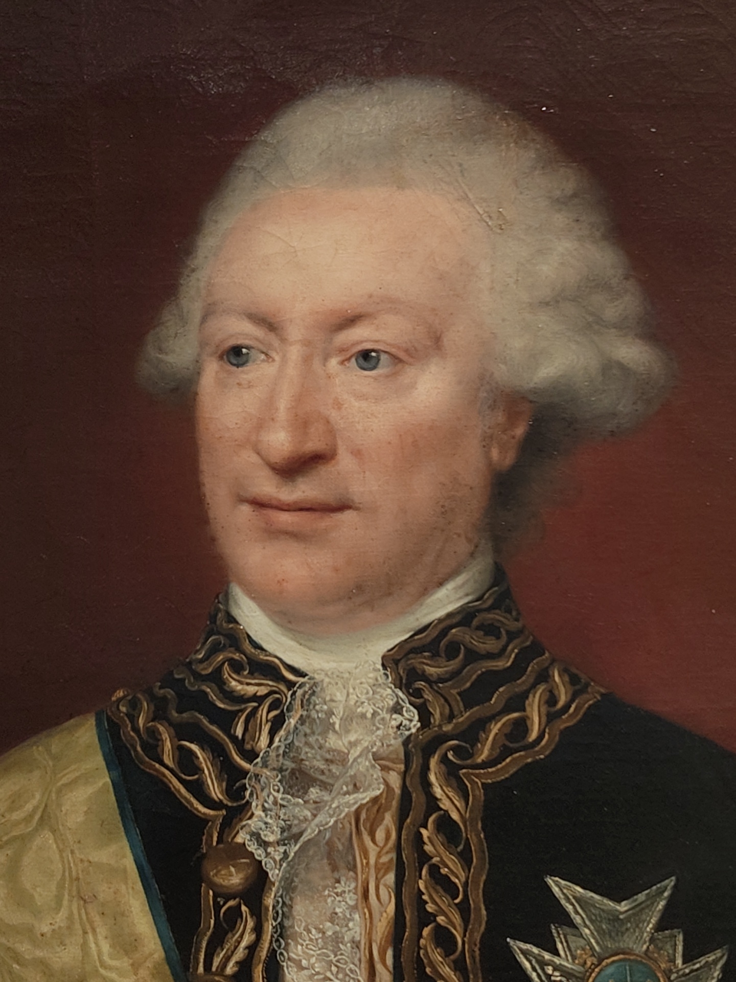 A portrait painting features a man with fair skin, blue eyes, and a wig of white, styled hair. He is dressed in a dark jacket with ornate gold detailing along the edges, complemented by a lace collar. A yellow sash with a blue stripe crosses his chest. A medal or badge is visible on his left side. The background is a solid, deep red color. The location of the image within the city of Genoa, Italy is unknown.