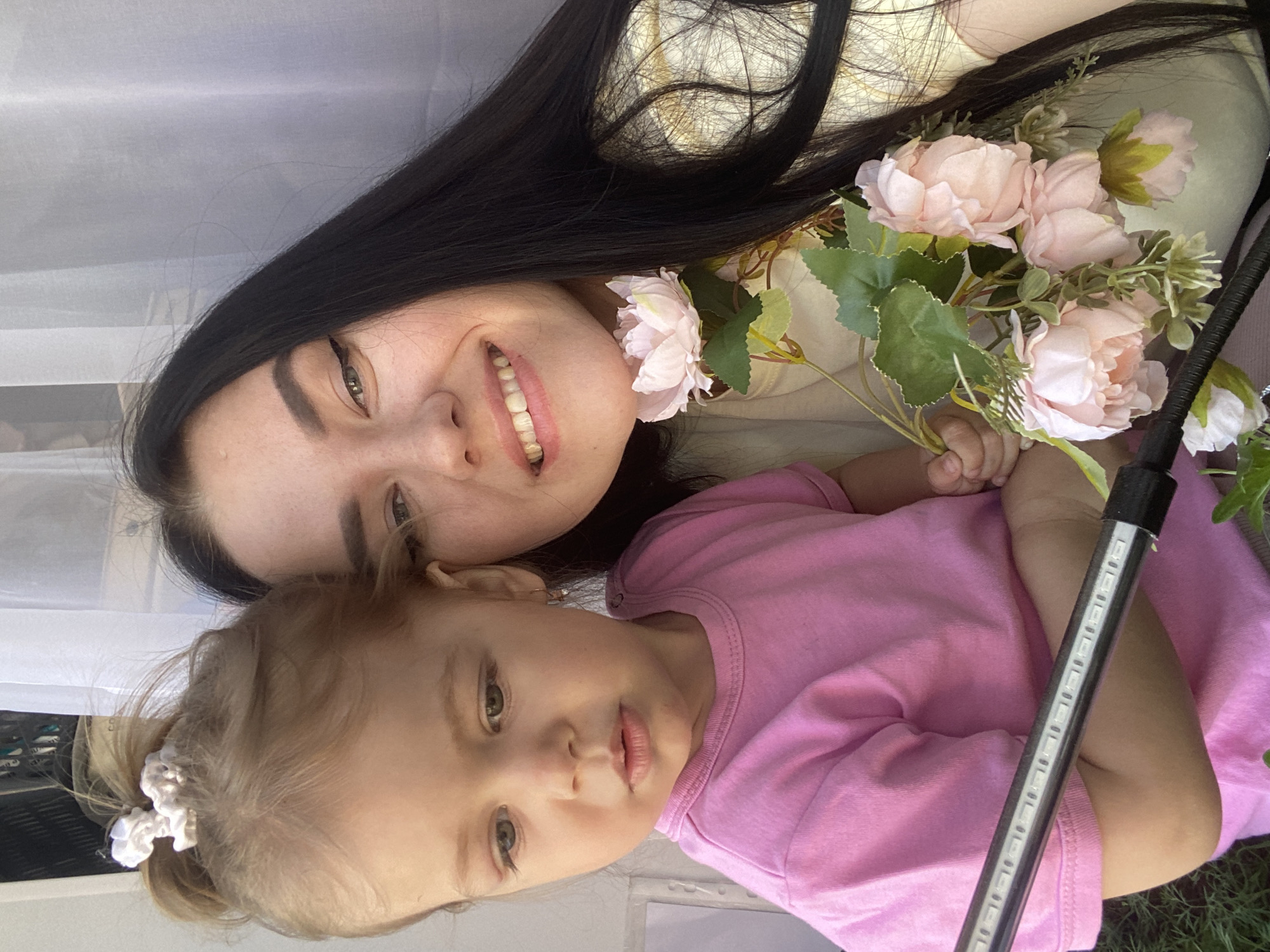A woman with long black hair and fair skin smiles, showing her teeth, next to a toddler with light brown hair pulled back into a ponytail with a white scrunchie. The toddler wears a pink shirt and gazes at the camera. The woman holds a bouquet of light pink flowers with green leaves. A black pole with white lights is near the toddler. The scene appears to be inside, with a white curtain in the background. Based on the location tag, the scene is in Chaikovsky, Russia.