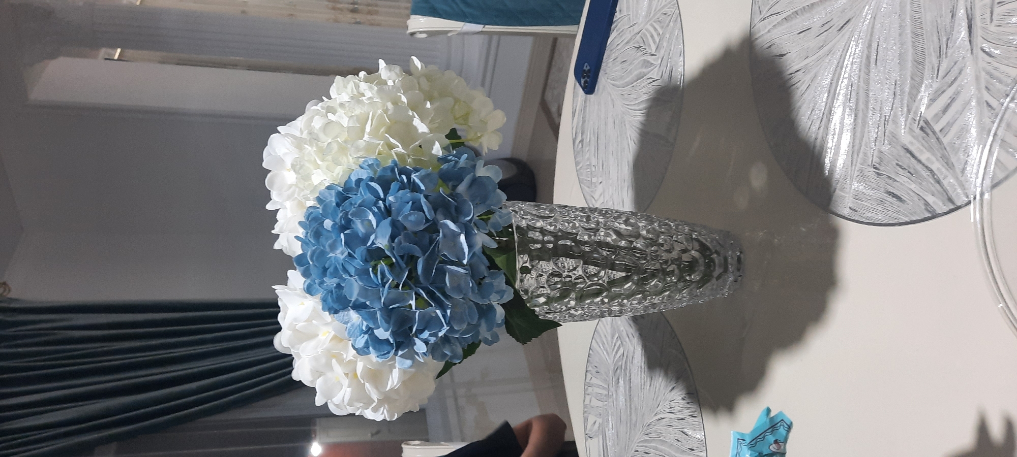 A close-up, vertical shot focuses on a glass vase filled with artificial hydrangeas. The vase, textured with a bubble-like pattern, is positioned on a round, light-colored table. The hydrangeas are arranged in a cluster, with blue blooms at the forefront and white blooms forming the outer layers. Two ornate, circular placemats are visible on the table, one beneath the vase and another to the right. A blue smartphone rests on the placemat to the right. In the background, dark teal curtains hang on the left side of the frame. To the right of the curtains, a white column is visible, and beyond that, a hint of patterned drapery. A person's arm and shoulder, clad in a dark shirt, are visible on the far left, partially obscuring the scene. The lighting suggests an indoor setting, possibly during the evening. There is no visible indication or context suggesting this scene is located in Gudermes, Russia.