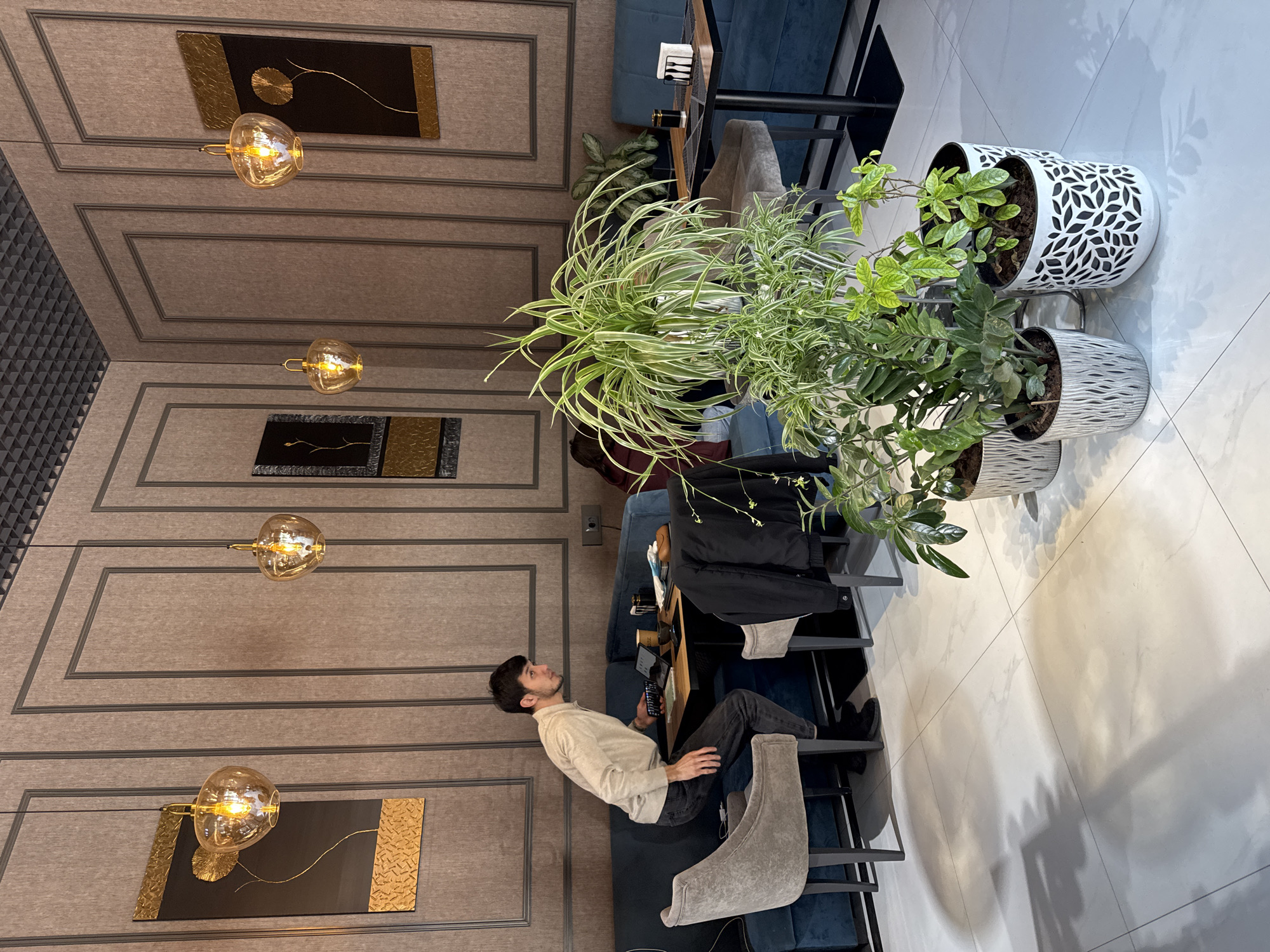 The image shows a man sitting at a table in a cafe, presumably in Tashkent, Uzbekistan. He is looking upwards and appears to be holding a remote or similar device. He's wearing a light-colored sweater and dark jeans. The cafe has a modern design with paneled walls, decorative artwork, and several hanging pendant lights. A variety of potted plants are placed near the seating area. The cafe has a clean, tiled floor and uses a combination of chairs and upholstered benches for seating.