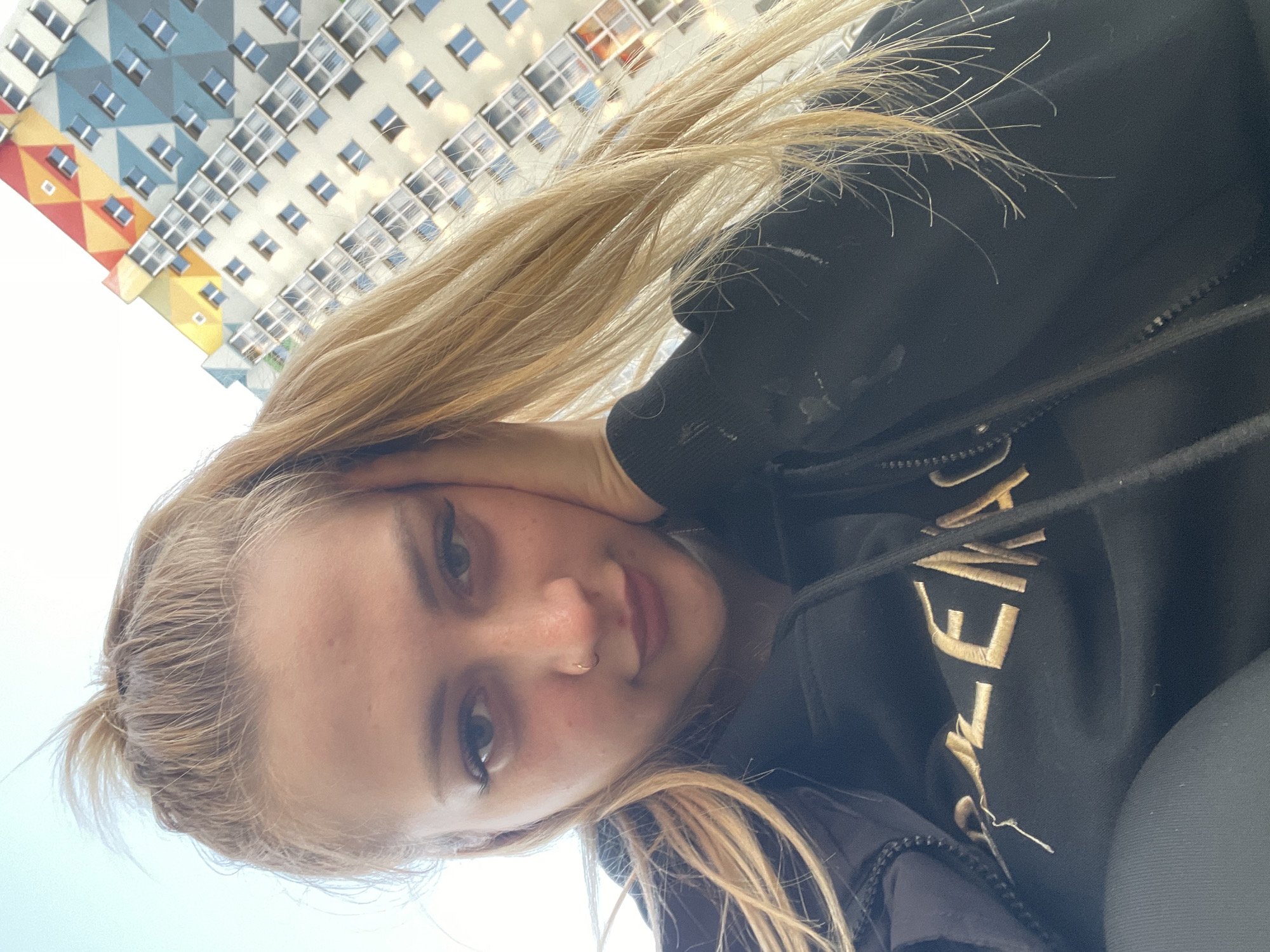 A blonde woman wearing a black zip-up hoodie with gold writing on the front poses for a selfie outdoors. Her hair is pulled back loosely. In the background is a high-rise building with a colorful, patterned facade, which identifies the location as Izhevsk, Russia. The woman is touching her face with one hand.