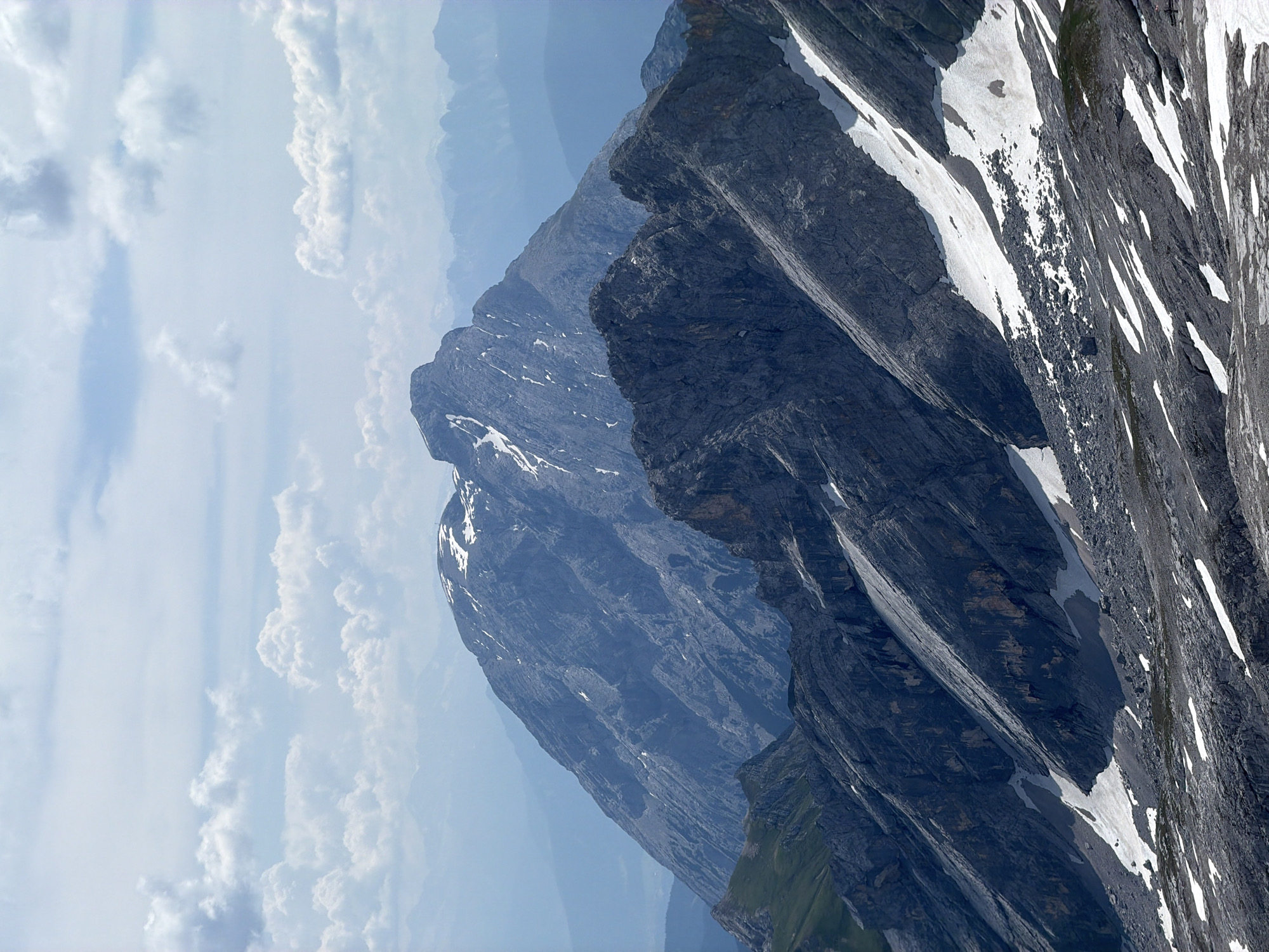 The image depicts a mountainous landscape with multiple layers of peaks under a cloudy sky. In the foreground, rocky slopes are dotted with patches of snow. The middle ground features darker, jagged mountain ridges with significant snow cover, and a green grassy slope on the left. The background is dominated by a large, imposing mountain peak with lighter grey rock and streaks of snow, appearing softer and more distant due to atmospheric haze. Further in the distance, more mountain ranges fade into a hazy blue. The scene suggests a high-altitude environment, likely within the Alps. This view is characteristic of the Zugspitze massif, Germany's highest peak, located near Garmisch-Partenkirchen. No people or specific man-made objects are clearly visible, with the exception of what appears to be a small structure or antenna on a distant slope. The overall impression is one of natural grandeur and rugged, remote terrain.